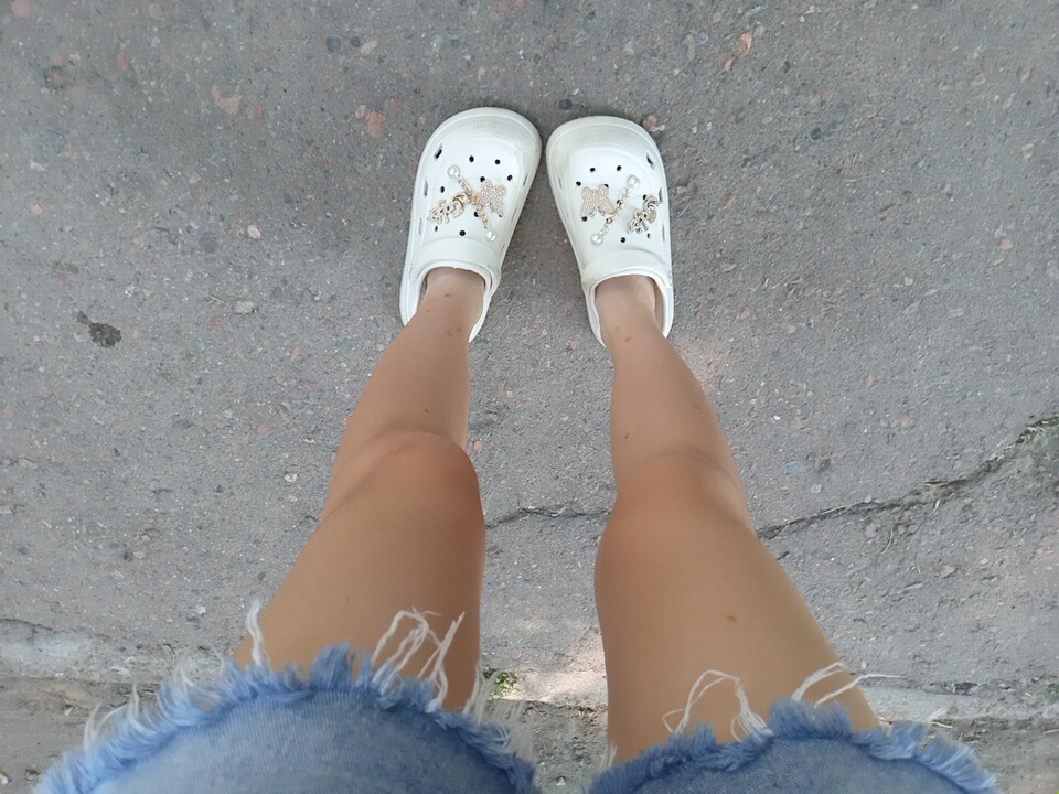 A person stands on an asphalt surface, possibly a sidewalk or road, in Angren, Uzbekistan. They are wearing a blue denim skirt with frayed edges and white Crocs. The Crocs are adorned with charms, including two butterflies and a charm that appears to spell out "Love". The lighting suggests it is daytime, with some shadows present. The background is blurred and appears to be an outdoor urban environment.