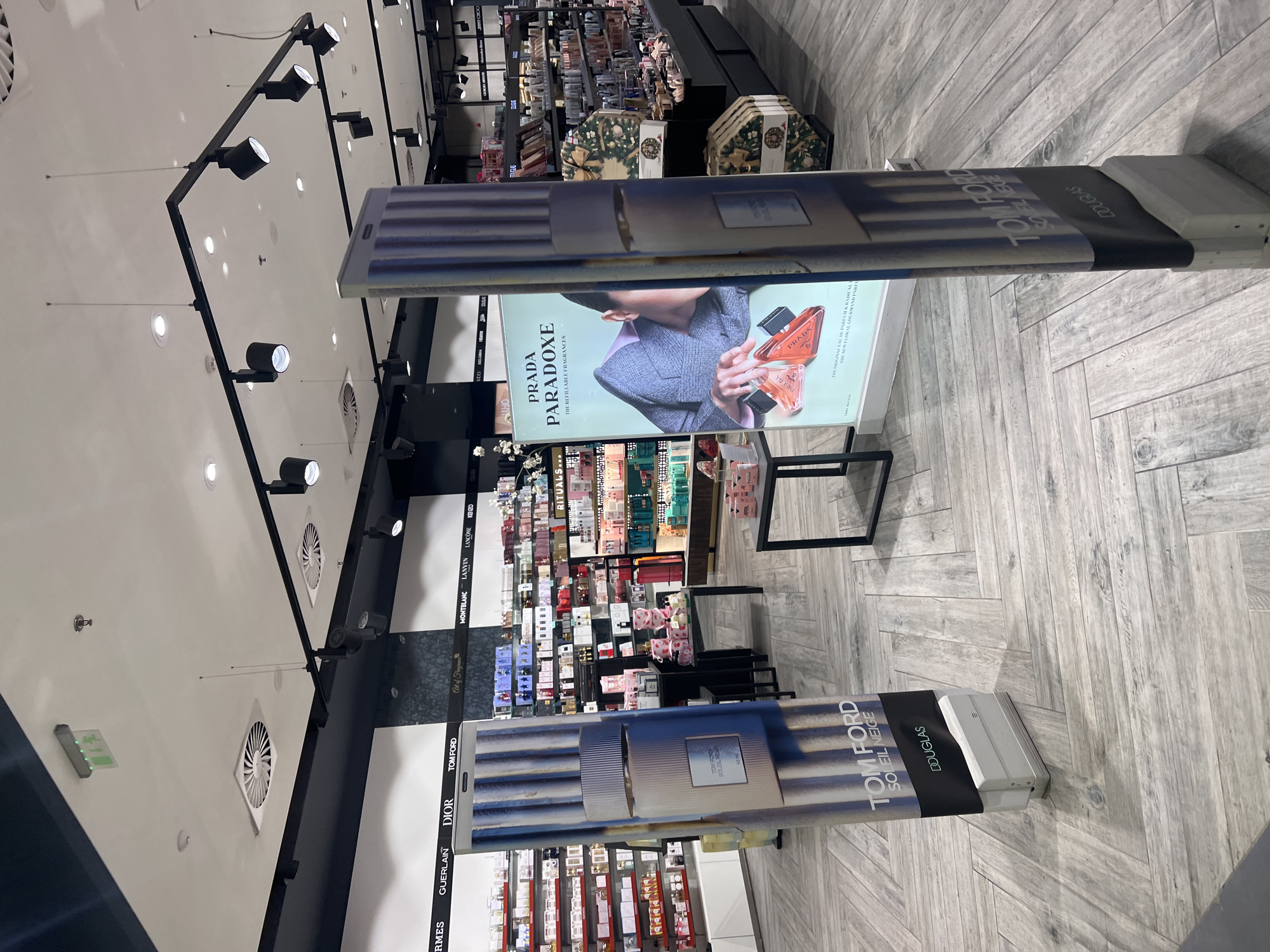 The image depicts the interior of a department store, likely a perfume or cosmetics section, with displays of various brands such as "Tom Ford," "Dior," "Guerlain," and "Prada". The displays feature promotional images and signage for the products. The foreground features two prominent advertising panels with images of "Tom Ford Soleil Neige" and "Prada Paradoxe". The retail displays with products stretch into the distance. The flooring has a herringbone pattern. The ceiling features track lighting. The location is not specified.