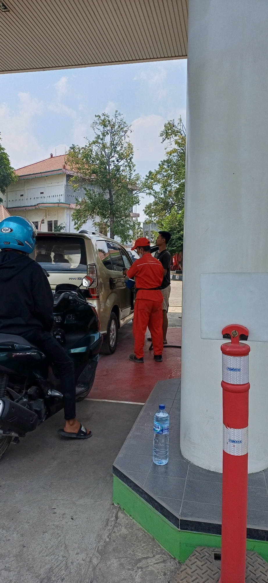 This image captures a scene at a gas station in Kecamatan Pati, Indonesia, likely during daylight hours under a partly cloudy sky. In the foreground, a person wearing a black hoodie and a blue helmet is seated on a dark green motorcycle. To their right, a gold-colored car is being refueled by an attendant dressed in a bright orange uniform, including a hat. The attendant is inserting a fuel nozzle into the car's gas tank. Another person, wearing a dark t-shirt and light-colored shorts, stands beside the car, facing the attendant. A bottle of "AQUA" brand water rests on a tiled surface to the right of the scene, near a red and white striped bollard. In the background, a multi-story building with a tiled roof is visible, surrounded by trees. The ground in the immediate vicinity of the refueling area is red, contrasting with the concrete and tiled surfaces elsewhere.