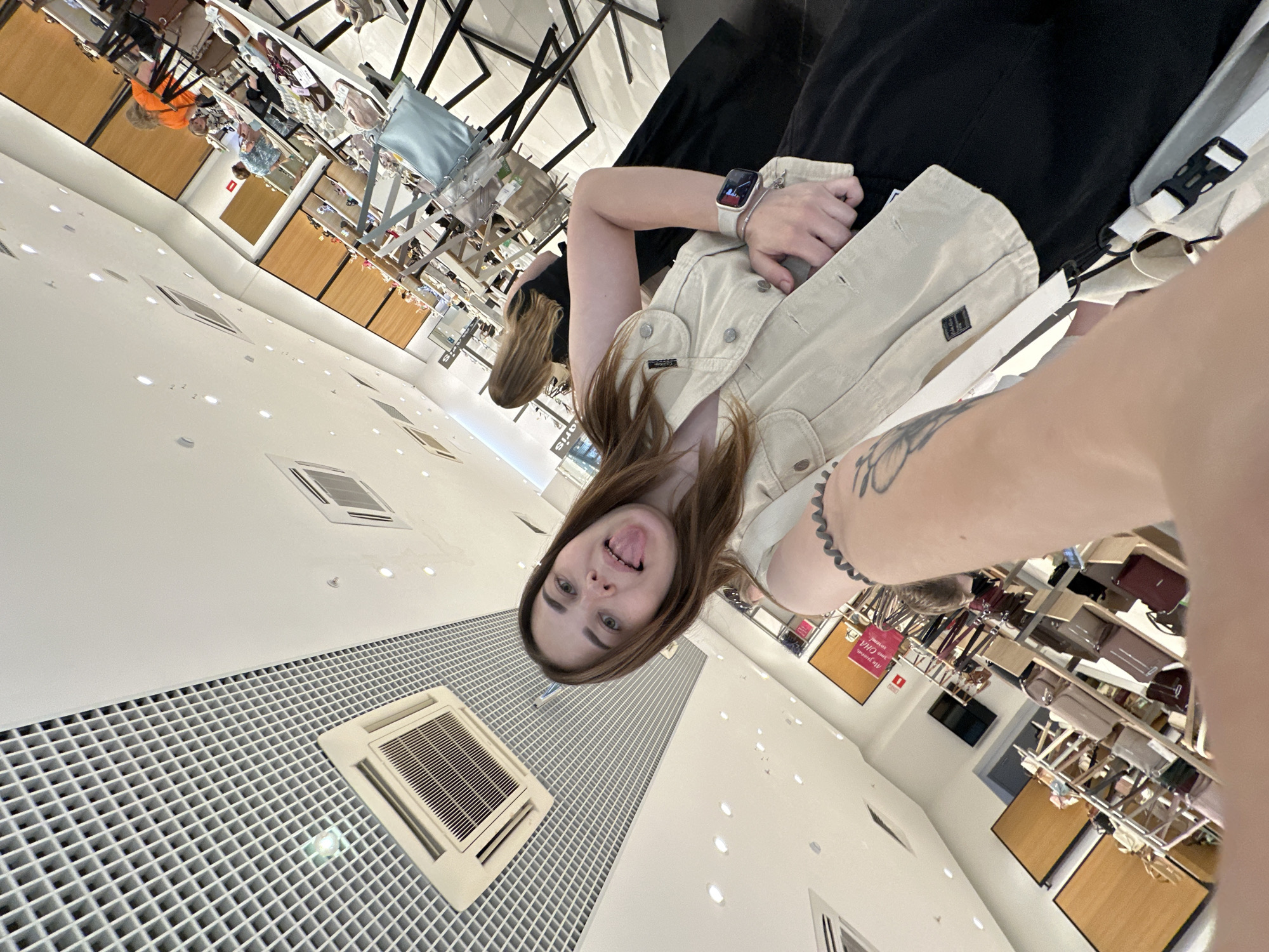 A selfie taken in the shopping gallery "Koltso" in Perm, Russia shows a person with long brown hair sticking out their tongue. They are wearing a light-colored vest over a black shirt. A wristwatch is on their left wrist. A tattoo is visible on their left forearm. In the background are displays of handbags and other merchandise. The ceiling has a grid pattern with air vents.