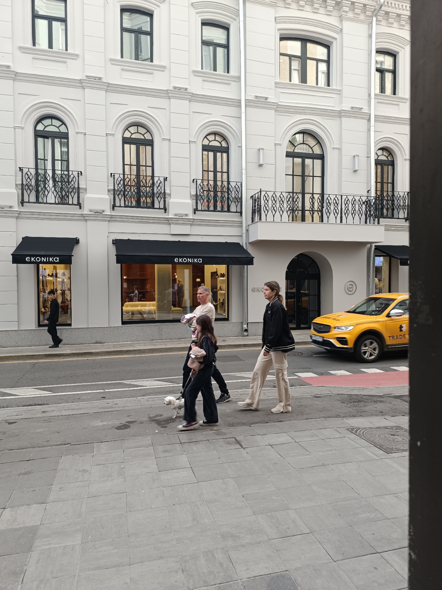 The image depicts a street scene in an urban environment. A white building with multiple windows and decorative architectural elements dominates the background. Below the building, there are two storefronts with black awnings and the word "EKONIKA" visible. A yellow taxi is driving on the street in front of the buildings.
In the foreground, three people are walking on a sidewalk. The person on the left appears to be a young man in dark clothing. To his right, a man and a young woman are walking together, and the woman is holding the leash of a small white dog. A younger girl is walking behind them, carrying a bag. The people are walking from right to left across the frame. The street has a crosswalk with red markings. The ground is paved with grey tiles.