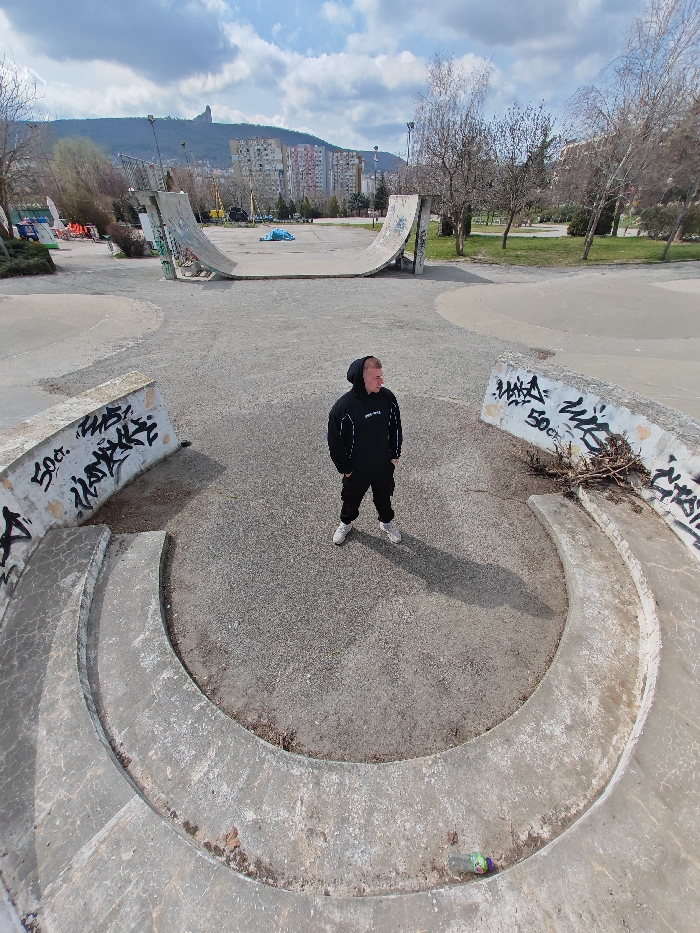 An outdoor skatepark scene in Shumen, Bulgaria, is depicted under a partly cloudy sky. A single male individual stands in the center of a circular concrete depression in the foreground. He wears a black hooded sweatshirt with white detailing, black pants, and light-colored athletic shoes, and is looking slightly upward and to his left. A clear plastic bottle with a purple and green cap lies within the concrete depression to his lower right. The skatepark features include a large concrete half-pipe in the midground and various concrete barriers, some adorned with black graffiti such as "50G." Beyond the skatepark, bare deciduous trees are visible, followed by multi-story residential buildings on a distant hillside. In the far background, a prominent, distinctive rock formation, consistent with the Shumen Plateau, rises against the sky. The scene is illuminated by direct sunlight, casting distinct shadows.