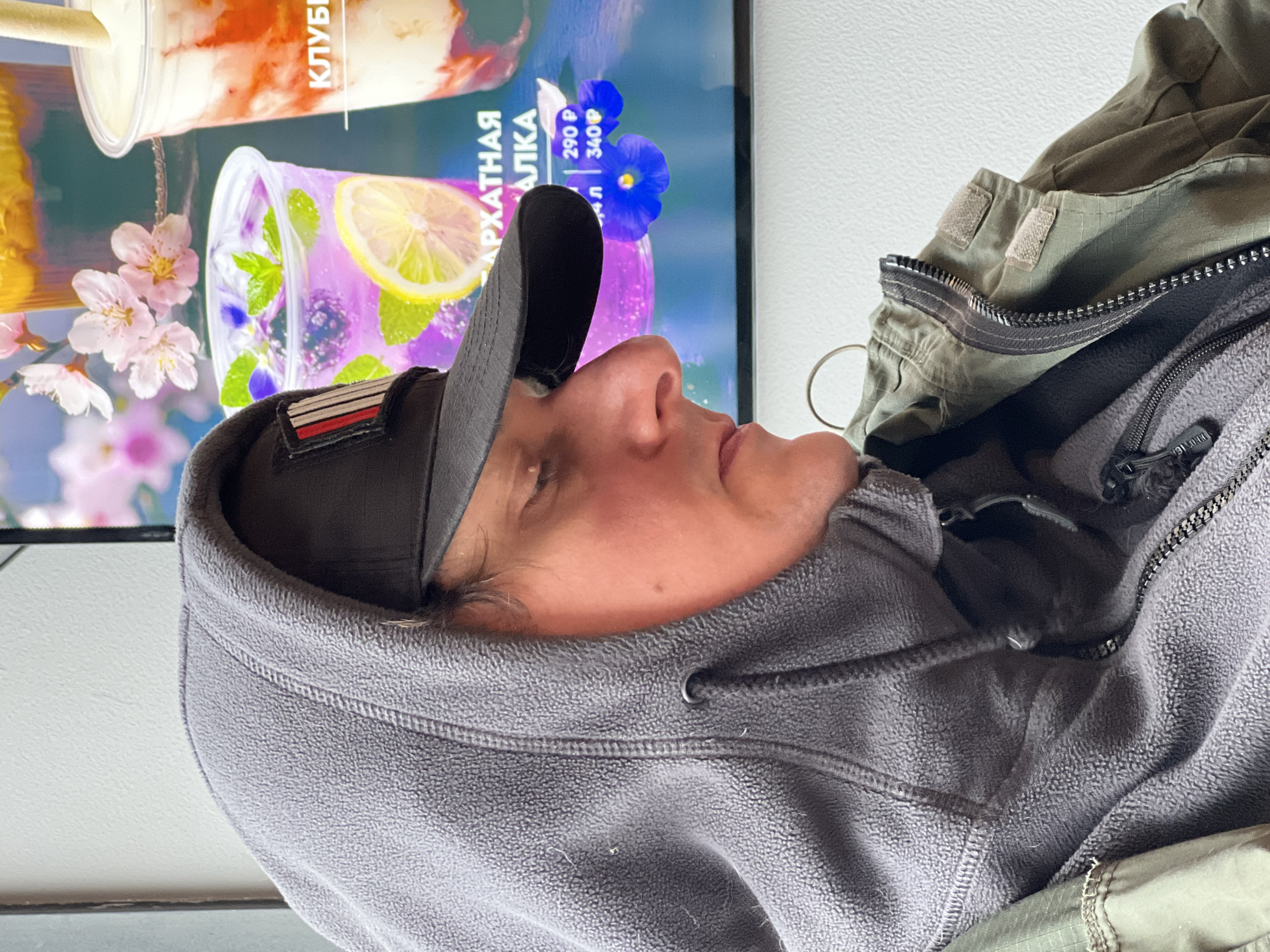 The image presents a close-up, side-profile view of a person within what appears to be a commercial establishment, possibly a cafe or fast-food place, located in Mytishchi, Russia. The person, an adult whose face is partially visible showing their nose, lips, and chin, is looking upwards and to the right, seemingly at a digital display in the background.
They are dressed in layered clothing, including a dark grey baseball cap featuring a distinctive rectangular patch with red, white, and black horizontal stripes on its side. Beneath the cap, they wear a grey fleece hoodie, and over that, an olive green utility-style jacket with visible zippers and velcro tabs.
The background is dominated by a brightly lit digital screen or advertisement board. This screen showcases two colorful beverages in clear plastic cups. One drink is a creamy, red-swirled concoction, likely strawberry-flavored, with partial Russian text "КЛУБН" (suggesting "Strawberry") visible above it. The second beverage is a vibrant purple-blue drink, garnished with fresh mint leaves, lemon slices, and dark berries. Above this drink, the Russian words "БАРХАТНАЯ" (Velvet) and "АЛКА" are visible. Prices are also displayed for the drinks in Russian Rubles: "290 Р" and "340 Р", with "0.4 л" indicating volume. The screen also features decorative elements like pink cherry blossoms on the left and a cluster of small blue flowers on the right.
The activity taking place is the person observing the menu or advertisement on the screen, possibly deciding on a drink. The clothing suggests a cooler indoor environment. The presence of Russian text on the display firmly places the scene in Russia, aligning with the provided location context of Mytishchi.