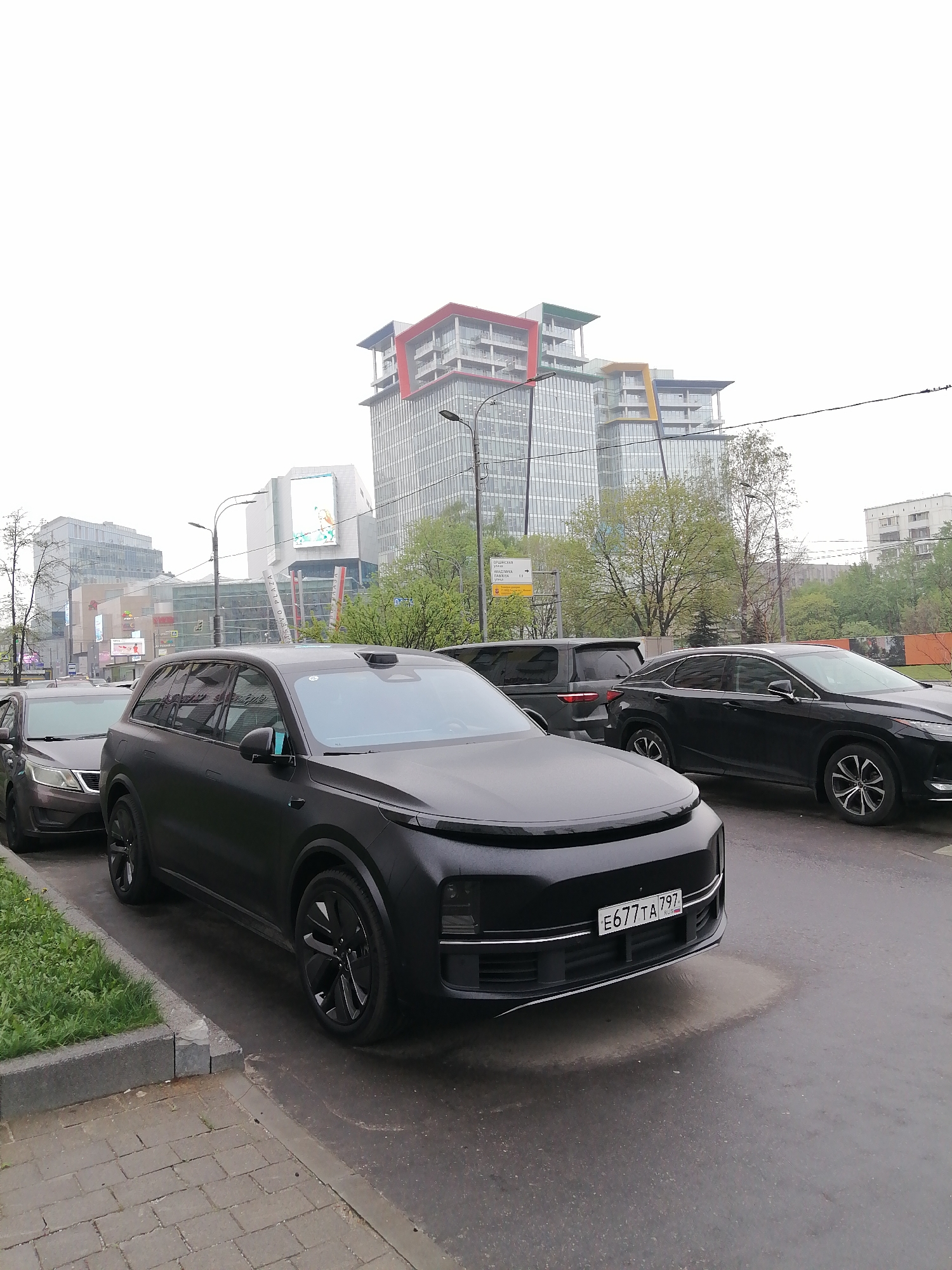 The image captures a street scene in Moscow, Russia. Dominating the foreground is a matte black Li Auto L9, a large SUV, parked on the side of a wet road. It is facing the camera and has a Russian license plate reading "E677TA 797." Next to it, a dark-colored car is partially visible. Further back, another black SUV and a black Lexus SUV are parked.
The background features modern architecture, including a tall building with colorful horizontal accents (red, green, yellow, blue) on its top floors. Other buildings with glass facades are also present, along with trees and streetlights. A billboard is visible in the distance. The sky is overcast, suggesting a cloudy day. The scene appears to be in an urban area, with a sidewalk visible on the left.