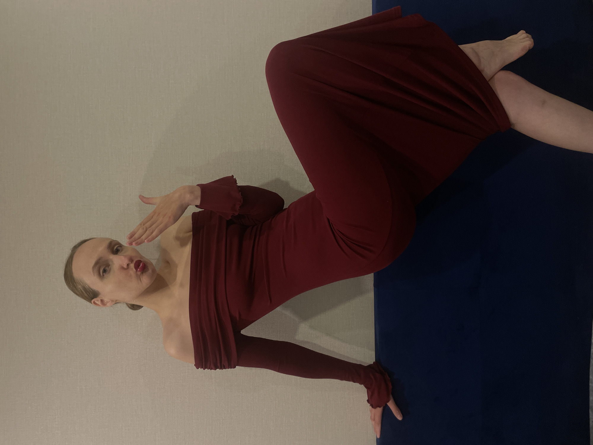 A woman in a dark red off-the-shoulder dress lies on a dark blue surface, possibly a mat or blanket. She is positioned against a neutral-colored wall. She has fair skin, her hair pulled back, and is blowing a kiss toward the viewer. Her dress has long sleeves with ruffled edges. The background appears to be a plain interior space. There is no direct evidence to confirm the location is in the city of Подшивалово, Russia.