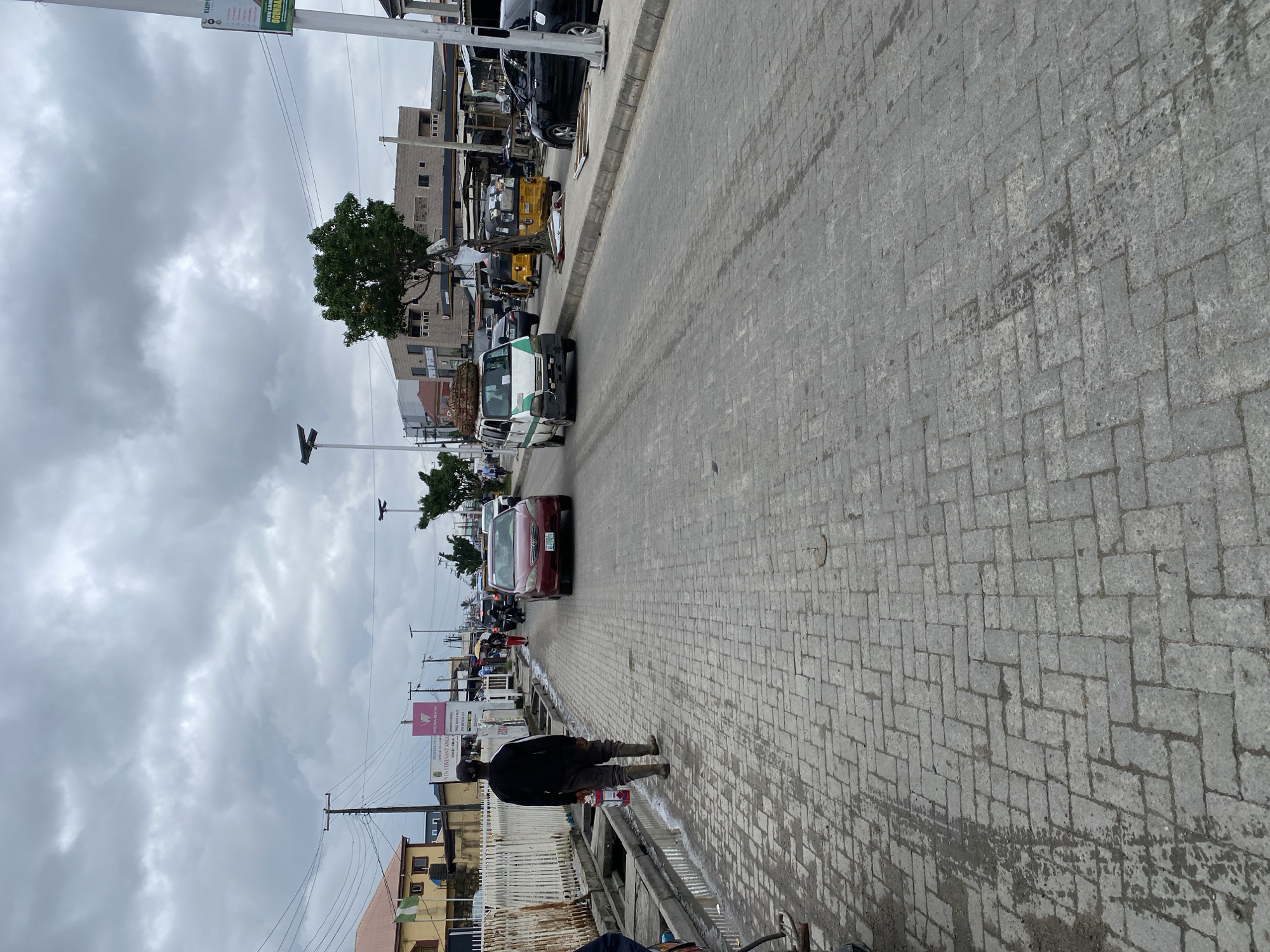 A cloudy, outdoor, daytime scene depicts a paved street with cobblestone-like blocks. In the foreground, a person walks away from the camera, carrying a red and gray cylindrical object. They are wearing dark clothing and tall boots. To the left, a white fence runs along the side of the street, with some drainage channels containing water. The street is populated with several vehicles, including a red car in the middle ground, a white and green van loaded with goods on top, and two yellow auto-rickshaws. Other vehicles and buildings are visible in the background, suggesting an urban environment. The sky is overcast with dense clouds.
