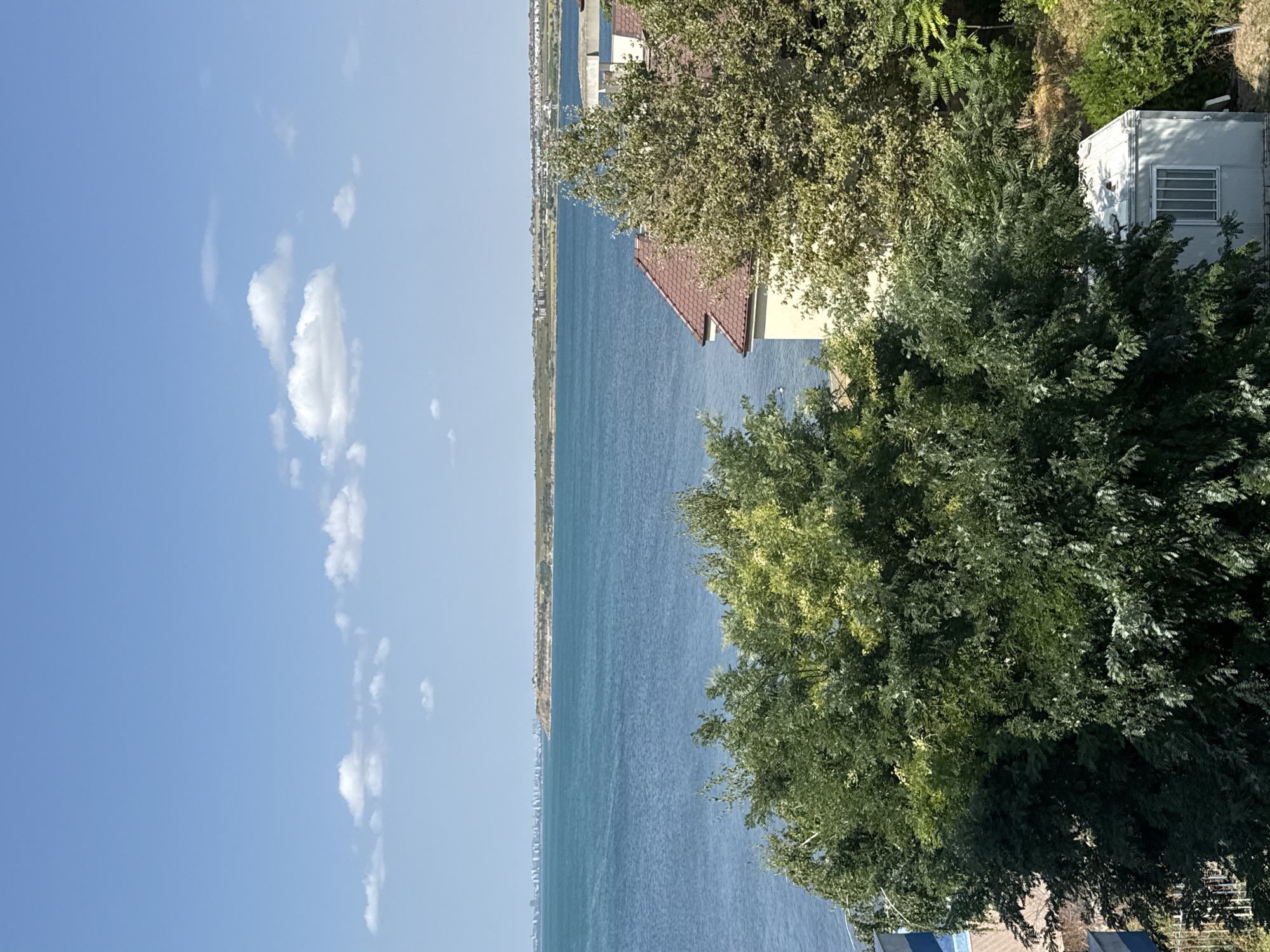The image captures a coastal scene in Pomorie, Bulgaria, on a bright, clear day with a blue sky dotted with scattered white clouds. The foreground is dominated by lush green trees, with some foliage in lighter, yellow-green hues. Partially visible behind the trees is a house with a reddish-brown tiled roof and beige walls. To the right, a small, white, box-like structure with barred windows is situated amidst the vegetation.
Beyond the trees and structures lies a wide expanse of calm, turquoise water, likely the Black Sea. In the distance, a long, low-lying coastline is visible. Along this distant shore, a city skyline with numerous buildings stretches across the horizon, interspersed with natural terrain. The overall impression is one of a peaceful, sunny day by the sea, with a blend of natural landscape and urban development.