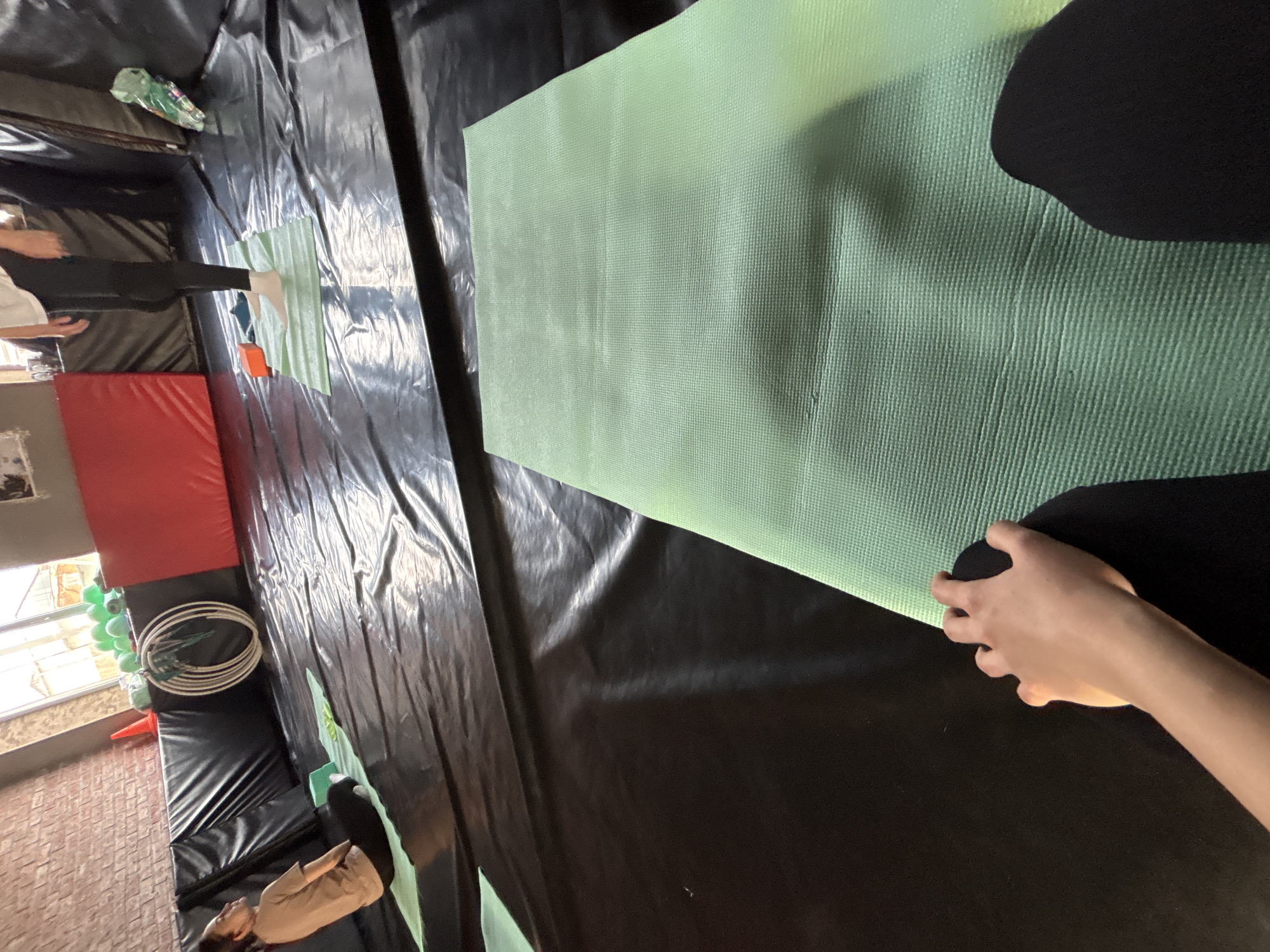 The image shows a person holding a green yoga mat in what appears to be an indoor activity center. The mat has a textured surface. The person is wearing black pants.
The background consists of black tarps, and in the periphery, a brick wall, windows, red pads, white hoops, and green balloons are visible. Other people are visible in the distance, walking by.
The image shows the act of setting up a yoga mat in an indoor activity center. The location is Ферма, Russia.