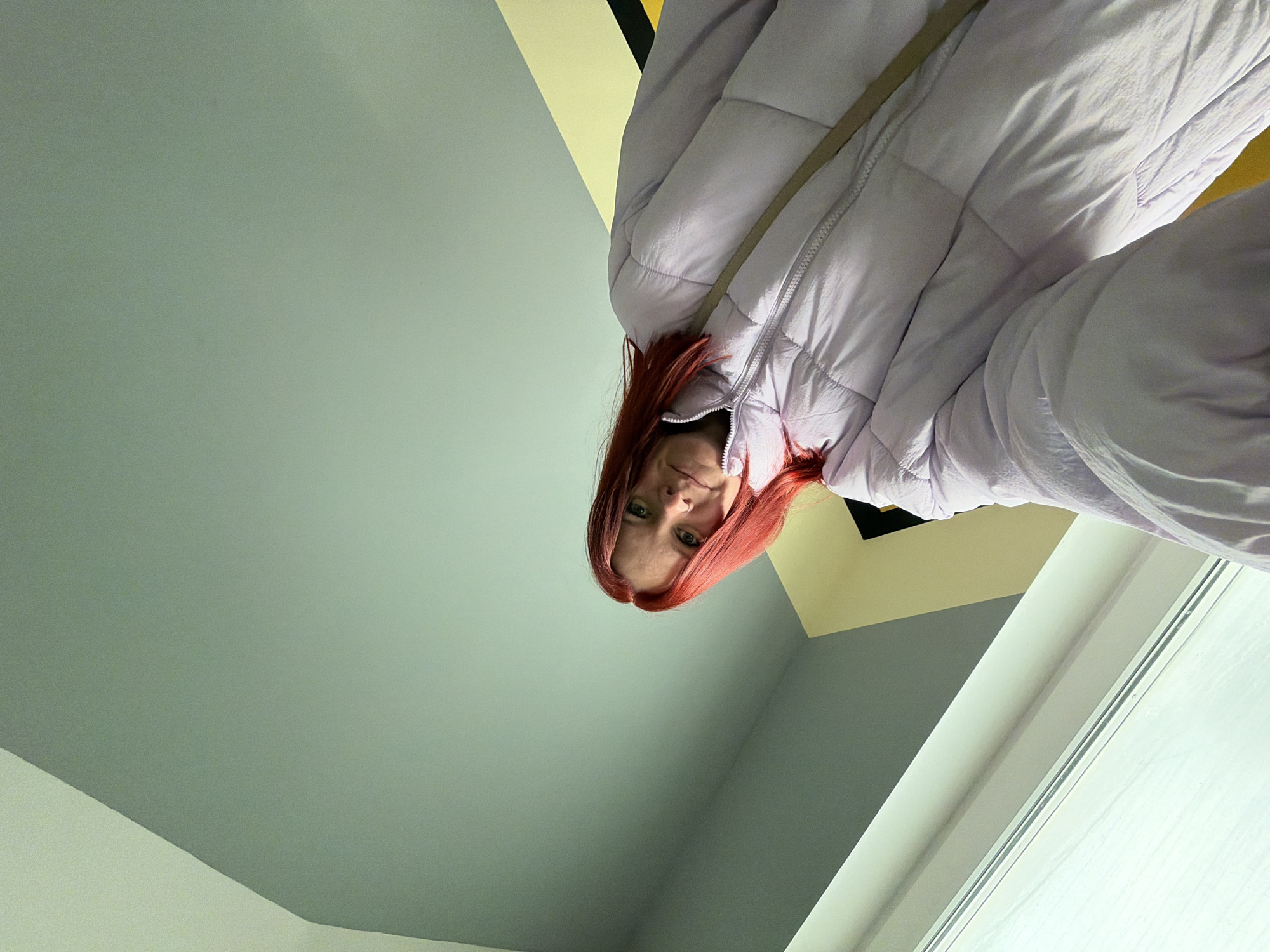A female individual with bright red hair and a nose ring on her left nostril is depicted in a low-angle selfie, looking directly at the camera with a slight smile. She is wearing a light purple puffer jacket with a visible zipper and a light-colored strap diagonally across her body, likely from a bag. The background is an interior space featuring a large, rectangular window with a white frame in the upper left, admitting diffused light. The interior walls are distinctly colored: a light greenish-blue wall occupies the upper right portion, transitioning to a light yellow section below, separated by a narrow black line. The specific location within Chaikovsky, Russia, cannot be determined from the visual evidence presented in the image.