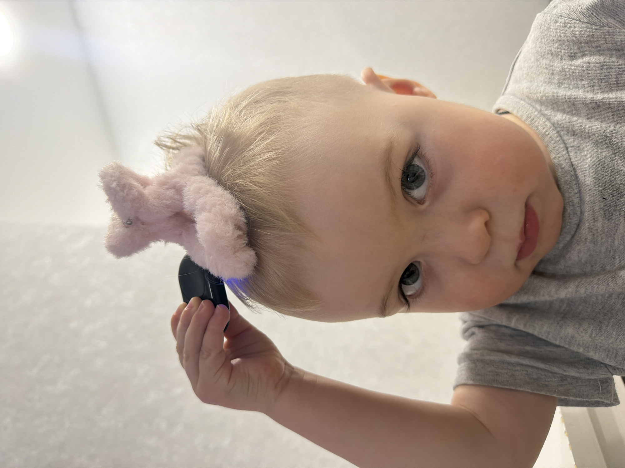 A young child, likely a baby or toddler, is captured in a close-up shot from a low-angle perspective, looking down and directly at the camera. The child has light blonde hair and light-colored eyes, possibly blue or green, with dark lashes. They are wearing a grey t-shirt.
The child holds a small, black, rectangular object in their right hand, which appears to emit a soft blue light onto a fluffy, pink plush toy. The plush toy, which resembles a small animal ear or a decorative element, is positioned on the side of the child's head, possibly as a hair accessory. The child's mouth is slightly open, revealing a hint of pink lips.
The background is predominantly bright white or very light grey, suggesting an indoor setting with a light-colored ceiling or wall above the child. The lighting is bright and even, indicating either daytime or a well-lit interior. There are no specific location cues visible within the image itself beyond a general indoor environment. No text is visible in the frame. The child's expression is neutral to curious, engaging directly with the viewer.