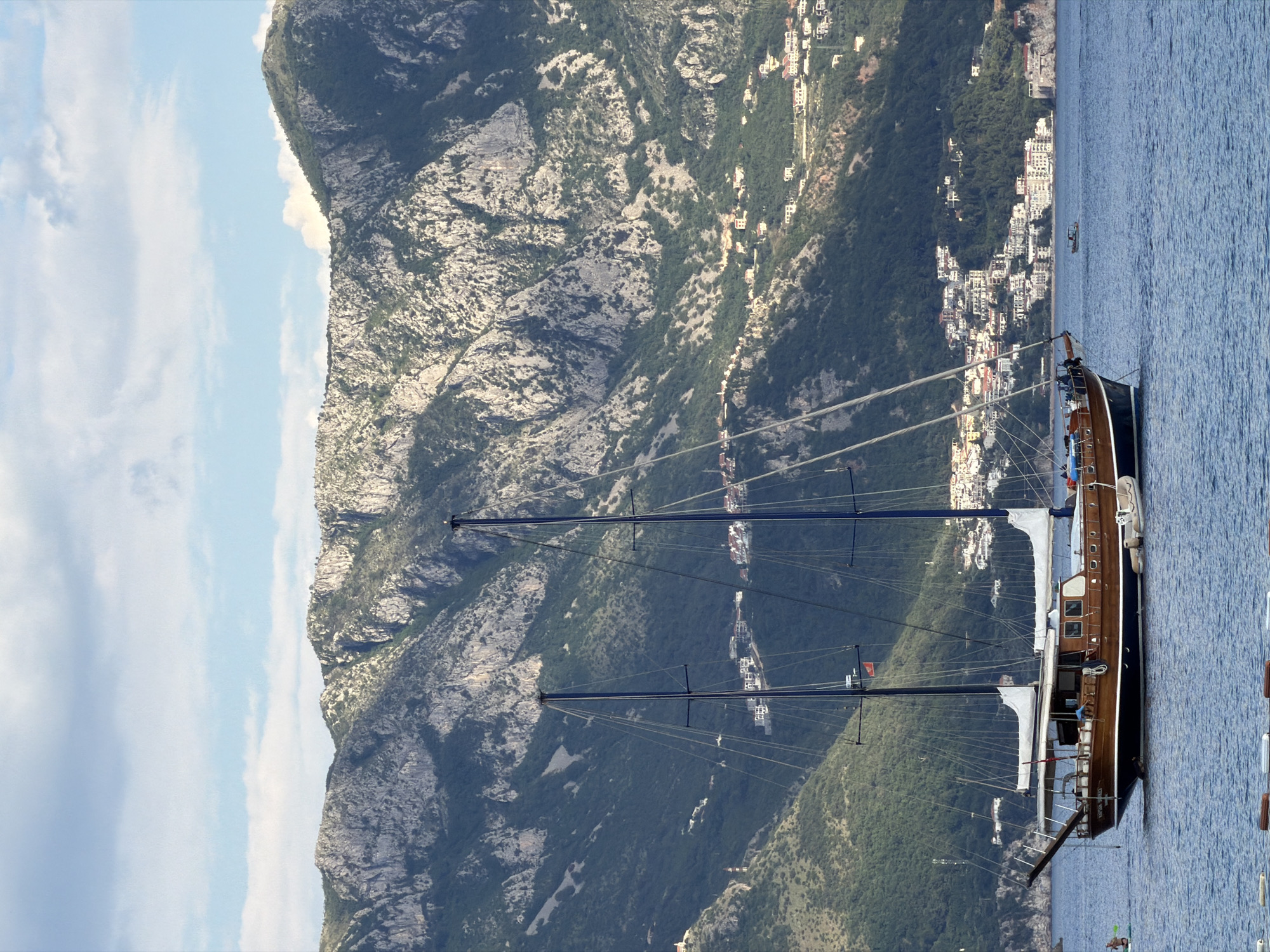 A high-angle shot captures a large sailboat moored in the Adriatic Sea near Budva, Montenegro. The dark blue sea contrasts with the tan and brown wooden deck and hull of the sailboat. Two tall masts rise from the deck, supporting thin cables. In the background, a steep mountain covered in green vegetation and gray rock formations stretches upward, with a cluster of white buildings dotting the lower slopes near the water's edge. The sky above is pale blue with scattered white clouds. The city of Budva is visible along the coastline, nestled between the mountains and the sea.