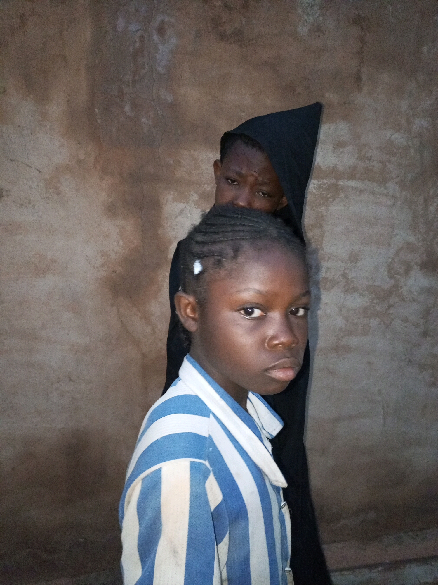 Two children are visible in front of a textured, light brown wall. A younger child, facing the camera, wears a blue and white striped shirt. Their dark hair is braided, and some white clips are visible. To the left, a second child is partially obscured, wearing a black garment that covers their head and shoulders. This child's eyes are visible, looking towards the camera. The lighting is dim, creating soft shadows. The image appears to be taken outdoors, possibly in a residential area of Tempure, Nigeria, given the style of the wall and the clothing of the subjects.