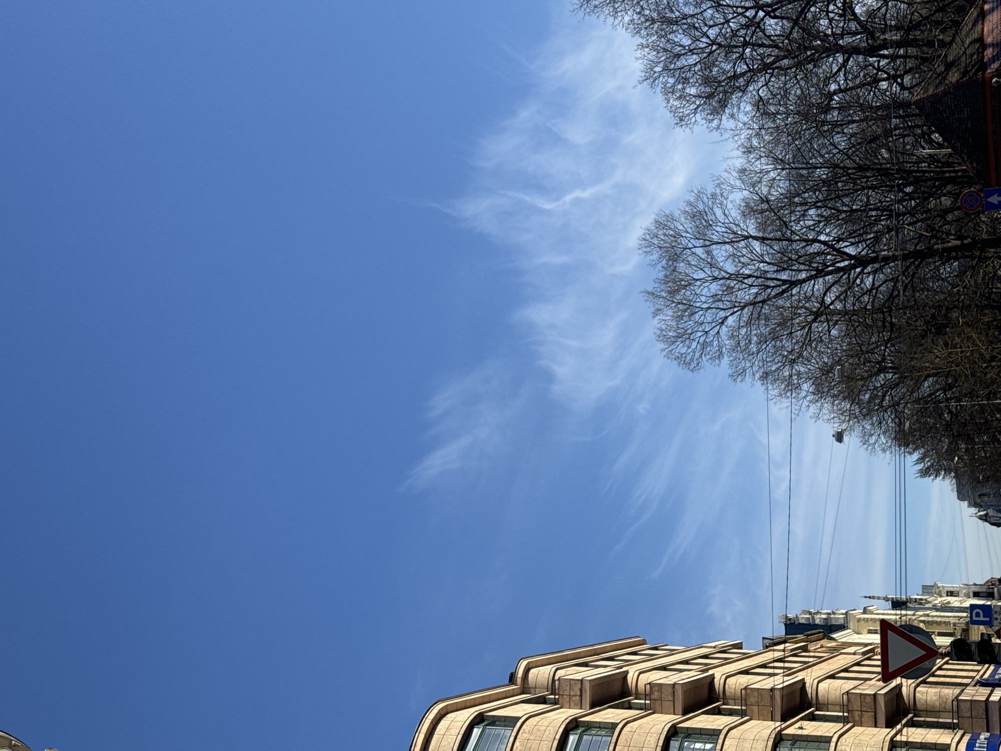 The image is taken looking up at the sky from a street in Riga, Latvia. The sky is a clear blue with wispy white clouds. Bare tree branches stretch across the top and right side of the frame. A building with a curving, layered facade occupies the lower right corner. Street signs are visible on the right, including a parking sign. The overall impression is of a bright, sunny day in an urban environment.