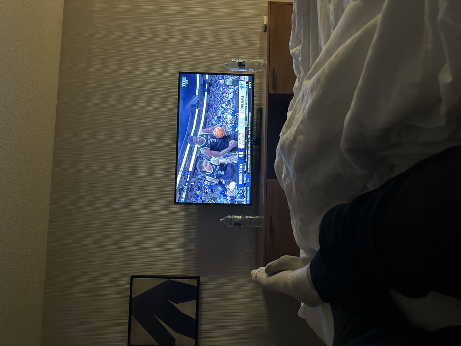 The image captures an intimate, relaxed scene, likely from a bedroom or hotel room, taken from the perspective of someone lounging in bed. The photo is rotated 90 degrees counter-clockwise from its natural orientation, showing a television screen as if it were vertical.
In the center of the frame, a flat-screen television is mounted on a wall featuring horizontal striped wallpaper. The TV is displaying a live basketball game. On the screen, two players in blue jerseys are visible on the court; one player (number 1) holds the basketball, while another player (number 5) is nearby. The scoreboard overlay indicates "SETON HALL 48" versus "GEORGETOWN 46" with "2:24" remaining in the "2ND HALF". A "BIG EAST" logo is prominently displayed, along with smaller text indicating "FOX" and "FS2" as the broadcast channel, and a "Program Alert".
Below the television, on what appears to be a narrow wooden shelf or console, two clear plastic water bottles are placed. Further down the wall, below the TV and the console, a dark-framed piece of art hangs, featuring an abstract blue shape against a lighter background. The wall to the far left of the TV has a different, more granular texture compared to the horizontally striped wallpaper.
To the right side of the image, a large, rumpled white duvet or blanket covers the majority of a bed. From beneath the duvet, the lower legs and feet of a person are visible, clad in white socks. Dark pants or clothing are also partially seen around the ankles. Part of a dark wooden furniture piece, possibly a headboard or nightstand, is discernible behind the white bedding. The overall lighting is subdued, suggesting a comfortable, indoor evening or night setting.