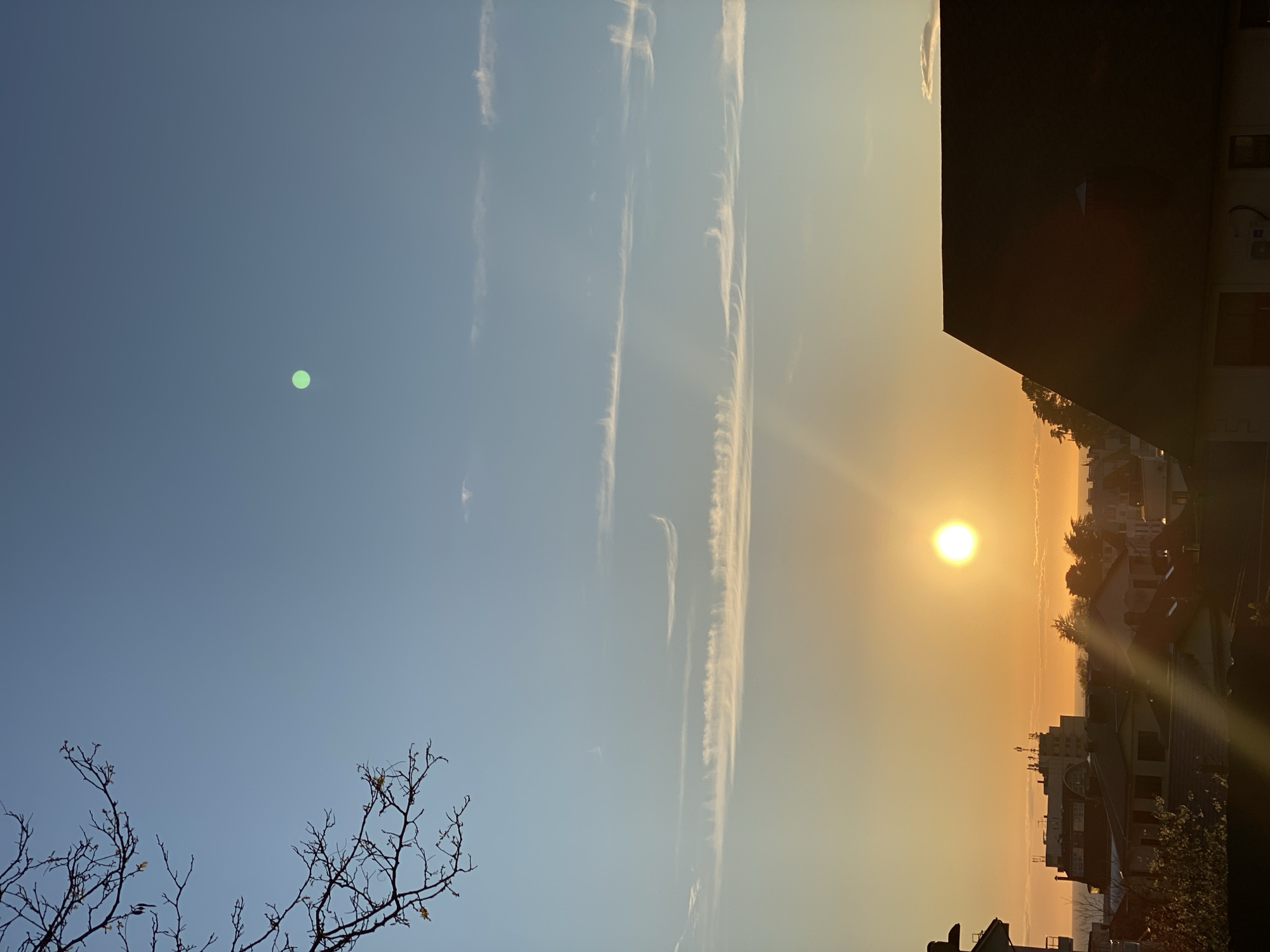 The image shows a sky at sunrise or sunset. The sky transitions from a clear blue on the left to an orange/yellow gradient on the right near the sun. There are thin, elongated clouds in the middle. A bare tree branch is visible in the lower left corner. On the right side of the image, rooftops and buildings are seen silhouetted against the bright sky. The location is purported to be Sochi, Russia, but there's no specific element in the image that confirms this location. There's a green dot that could be lens flair.