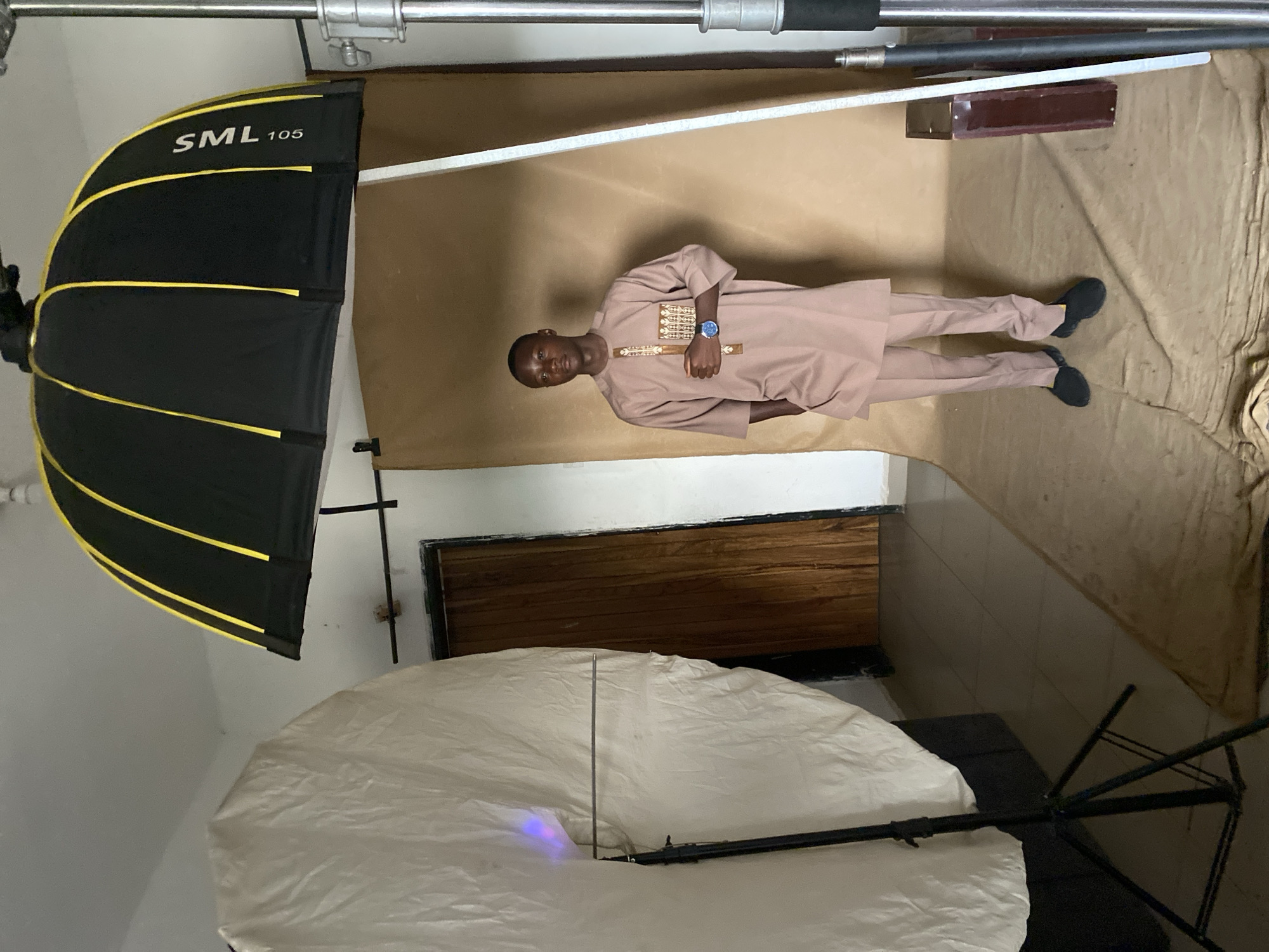 The image shows a man in Bukuru, Nigeria, standing in a photography studio. He is wearing a brown, traditional-style outfit and black shoes. The background is a beige backdrop. Lighting equipment, including a black and yellow softbox labeled "SML 105" and a larger white umbrella reflector, are positioned around the subject. The studio space has a tiled floor and a partially visible wooden door or panel in the background.