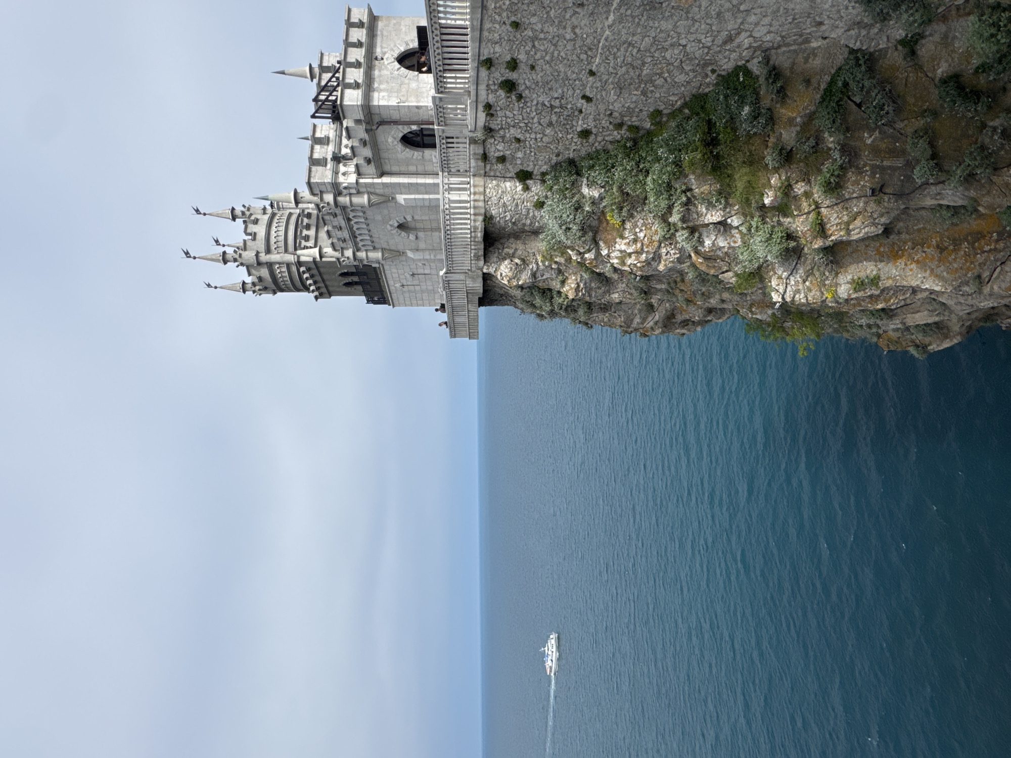 The image shows the Swallow's Nest castle perched on a cliff overlooking the Black Sea. The castle is built in a neo-Gothic style and is made of light-colored stone. The cliff is covered in vegetation and slopes down to the sea. The sea is a deep blue color and there is a boat visible in the distance. The sky is light blue and the castle is on the northern coast of the Crimean Peninsula near Yalta, in the city of Ливадийский поселковый совет, Ukraine.