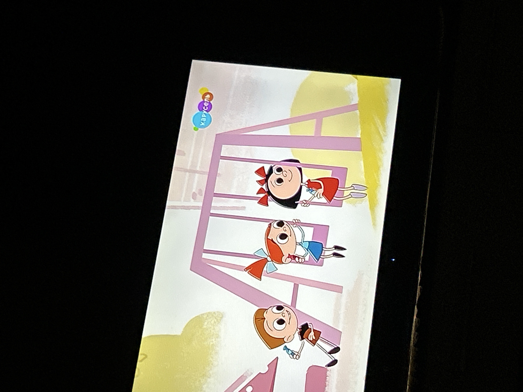 The image shows a television screen displaying a cartoon. Three animated characters are visible: a boy with brown hair and a red shirt, and two girls with red dresses and pigtails. One girl is on a pink swing, and the other is standing next to her. The boy is standing to the left of the girls, holding a blue object. In the background, there is a pink swing set and a yellow and white grassy area. The top right corner of the screen shows a logo with colorful circles and the word "Карусель" written in blue. The television is dark, suggesting it is not actively being used. The image is cropped to show only the screen and a small portion of the surrounding darkness. The location of Gelendzhik, Russia, is not discernible from the visual content of the image.