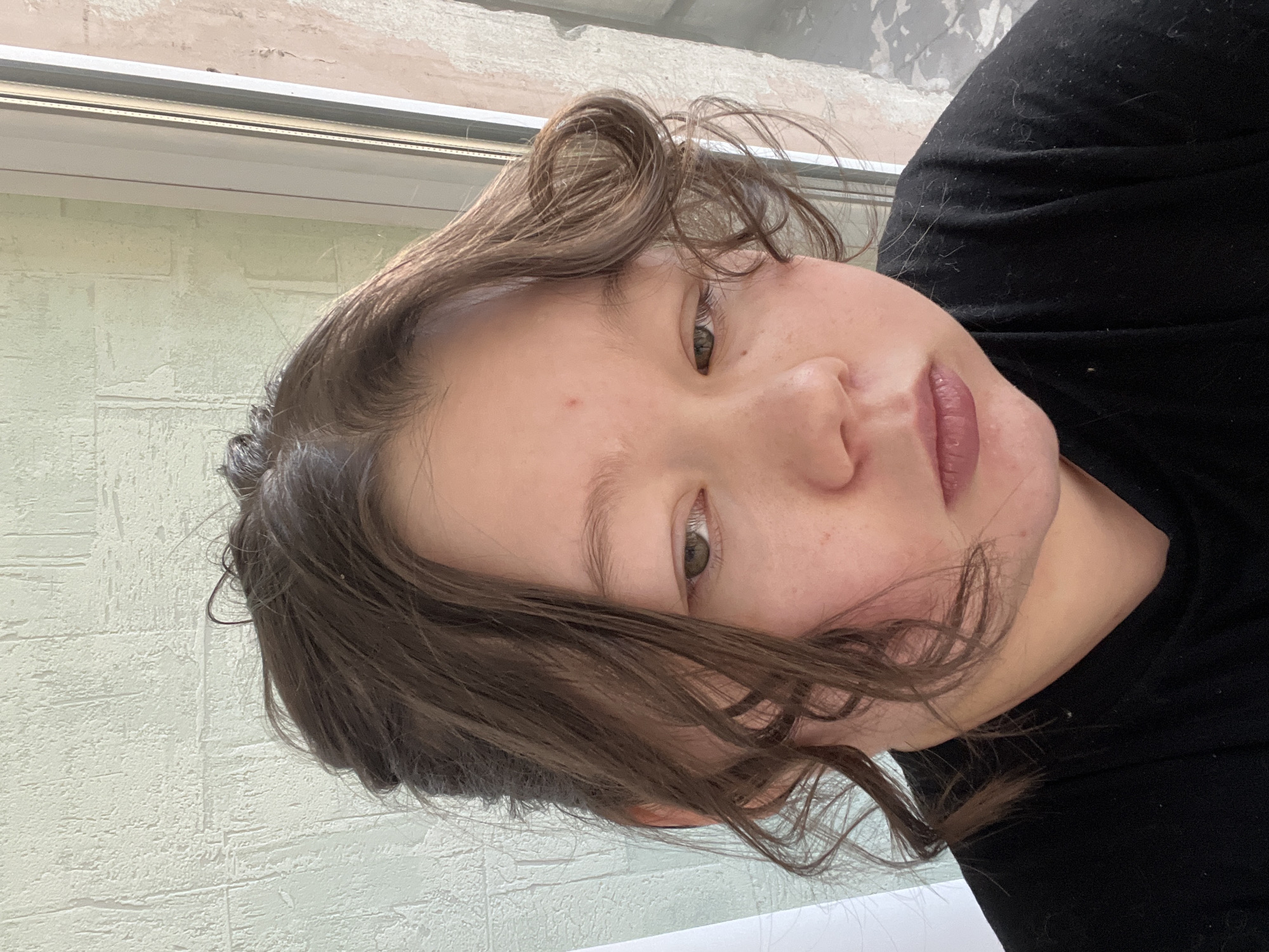 A close-up selfie of a person with light skin, brown wavy hair, and green eyes is presented. The person is wearing a black garment. The background includes a window with a white frame and a wall with a textured, light-colored surface. The image does not contain enough information to identify the location in Nizhny Tagil, Russia, or provide specific details about actions, interactions, or other elements.