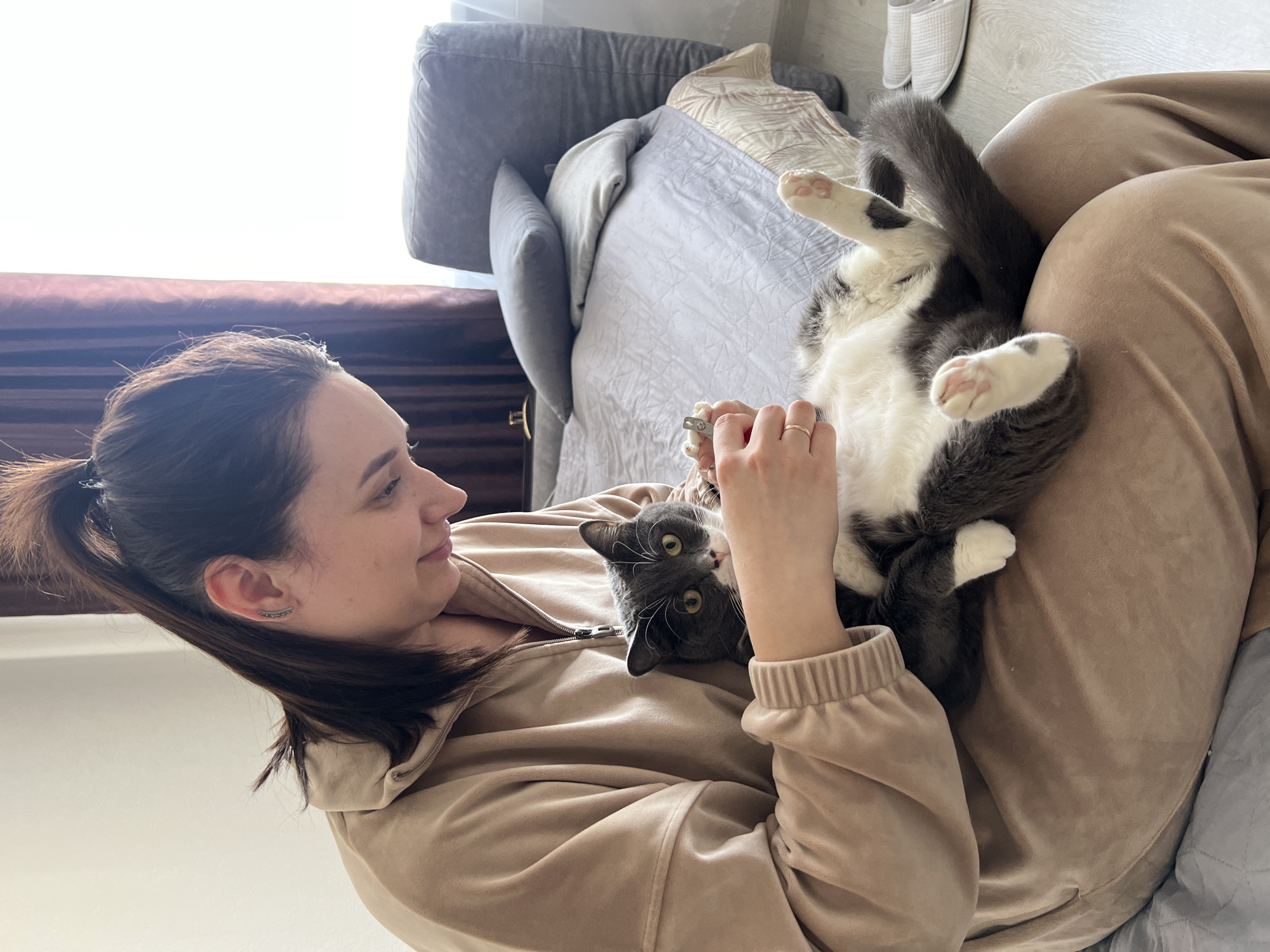 A woman with brown hair pulled back in a ponytail, wearing a beige velour tracksuit, sits on a bed and looks down at a gray and white cat lying on its back on her lap. The woman's right hand is holding a pair of nail clippers. The scene takes place indoors, with a couch and pillows in the background and a pair of slippers visible in the upper right corner. There are no obvious visual cues indicating the location of the image is Tyumen, Russia.