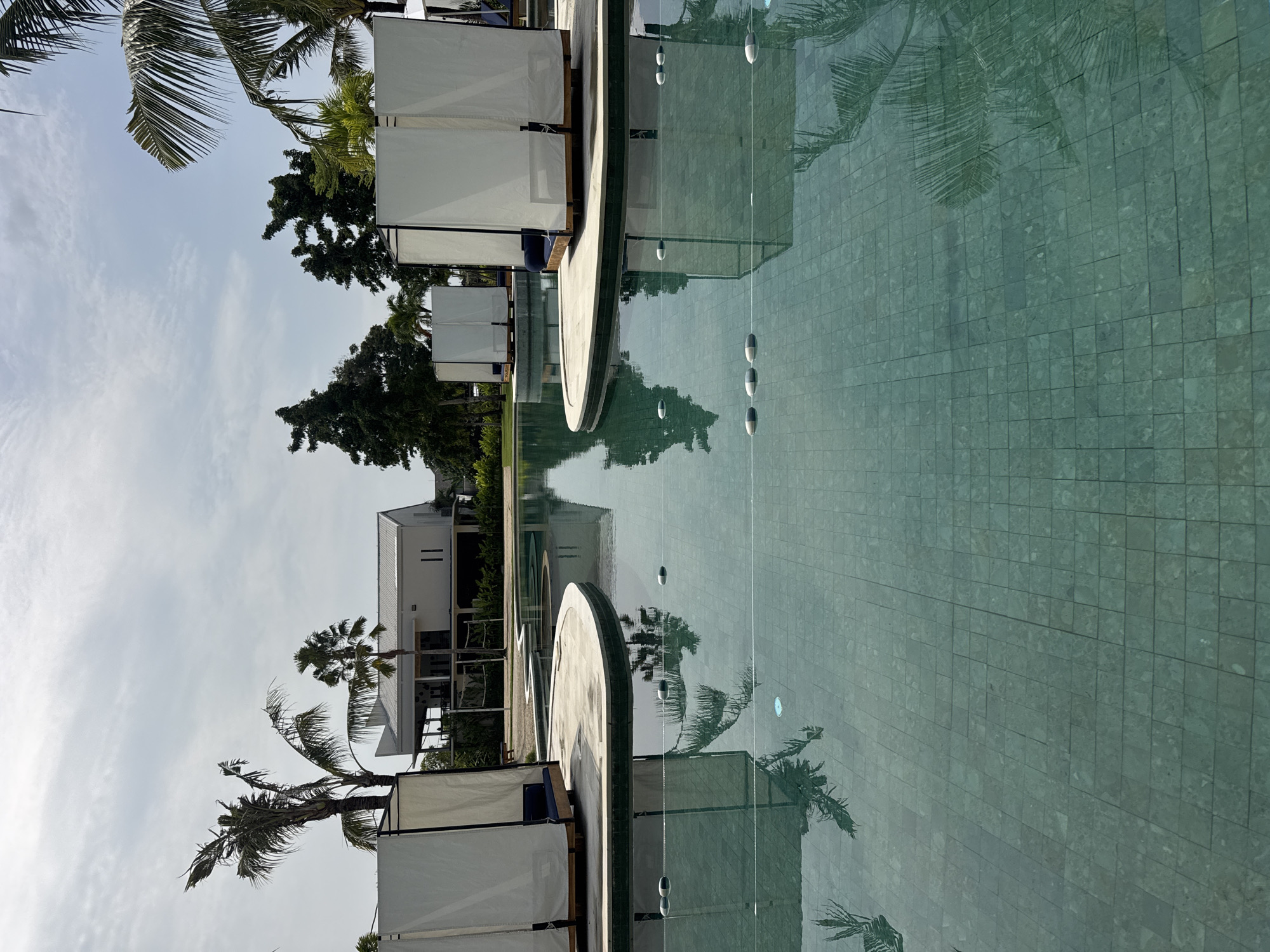 The scene depicts an outdoor, unoccupied swimming pool area located in Kuta, Indonesia. The expansive swimming pool is surfaced with teal-green square tiles, holding calm, reflective water that mirrors the sky and surrounding vegetation. Strategically placed within or adjacent to the pool are multiple open-sided cabanas, each constructed with a light-colored wooden base and white fabric sides, containing dark blue cushions. These cabanas rest on raised, light-grey concrete islands or platforms. Sections of pool divider ropes with white and grey cylindrical floats are visible on the water's surface. In the background, a modern, two-story white building with a distinctive sloped metal roof stands amidst dense tropical foliage, including numerous tall palm trees. A volleyball net is discernible in the grassy area between the pool and the building. The environment is illuminated by bright natural daylight, and no individuals, interactions, or specific actions are present within the frame.