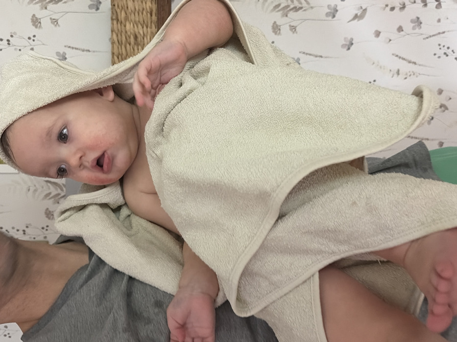 A baby, wrapped in a hooded towel, is held on a person's chest. The baby faces the left of the frame, eyes open and mouth slightly open. Redness appears on the baby's cheeks and around the mouth. The towel is light beige and covers most of the baby's body. The person's gray t-shirt is visible underneath. The background consists of a wallpaper with a floral pattern and a woven basket.