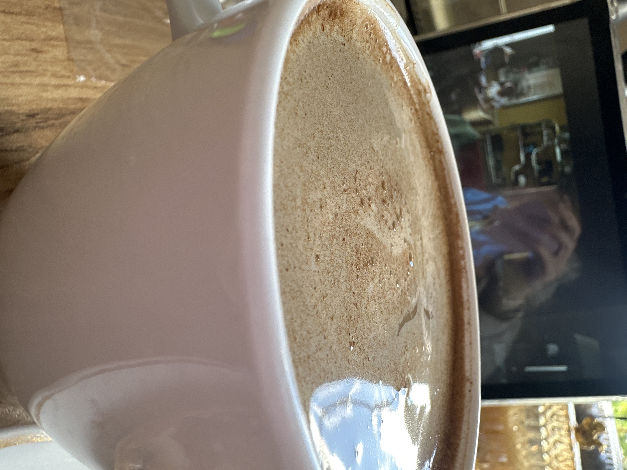 The image presents a close-up view of a white ceramic mug, which appears rotated 90 degrees clockwise, making it seem as if it's lying on its side. The mug is filled with a dark beverage, likely coffee, topped with a substantial layer of light brown foam or crema, the surface of which shows small bubbles and hints of reflectivity.
To the left of the mug, a light brown wooden surface provides a textured backdrop. In the background, a dark rectangular screen is prominently visible. This screen reflects a detailed image, including a person's hand and forearm, suggesting they are either interacting with the screen or holding the camera. Behind the reflection of the hand, complex shapes resembling pipes or industrial-style fixtures are discernible, likely part of the surrounding interior environment. Below the screen, further indistinct reflections show patterns.
The overall scene conveys an indoor setting in Stavropol, Russia. There is no visible text in the image, and no specific time of day or emotional context can be determined from the visual information.