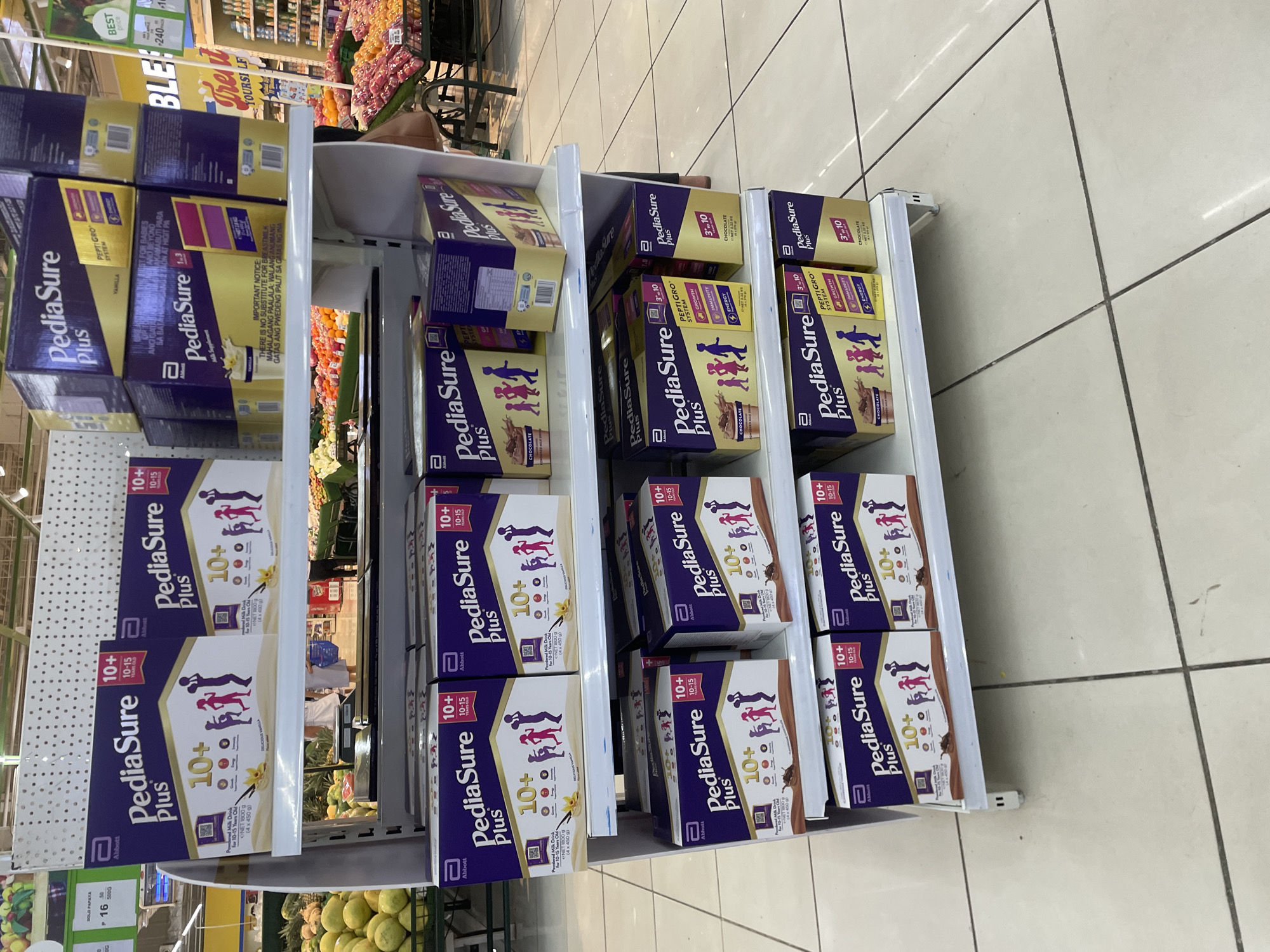 The image shows a retail store shelf stocked with PediaSure Plus milk supplement products. The products are arranged in multiple rows on white shelves. The packaging is predominantly purple and gold, featuring the PediaSure Plus logo, nutritional information, and illustrations of children. Some boxes indicate flavors such as "Delicious Vanilla" and "Chocolate."
The environment appears to be a supermarket or grocery store, with tiled flooring visible at the bottom of the image. Other products and store fixtures are partially visible in the background, including a display of fruits and vegetables to the left and a section with yellow signage to the right. The overall lighting suggests an indoor retail setting.
Based on common retail setups and signage that might be present in such stores, and given the context of Davao City, Philippines, this scene is likely set within a supermarket or hypermarket like SM Supermarket, NCCC Mall, or Robinsons Supermarket, all of which are prominent retail chains in Davao City.