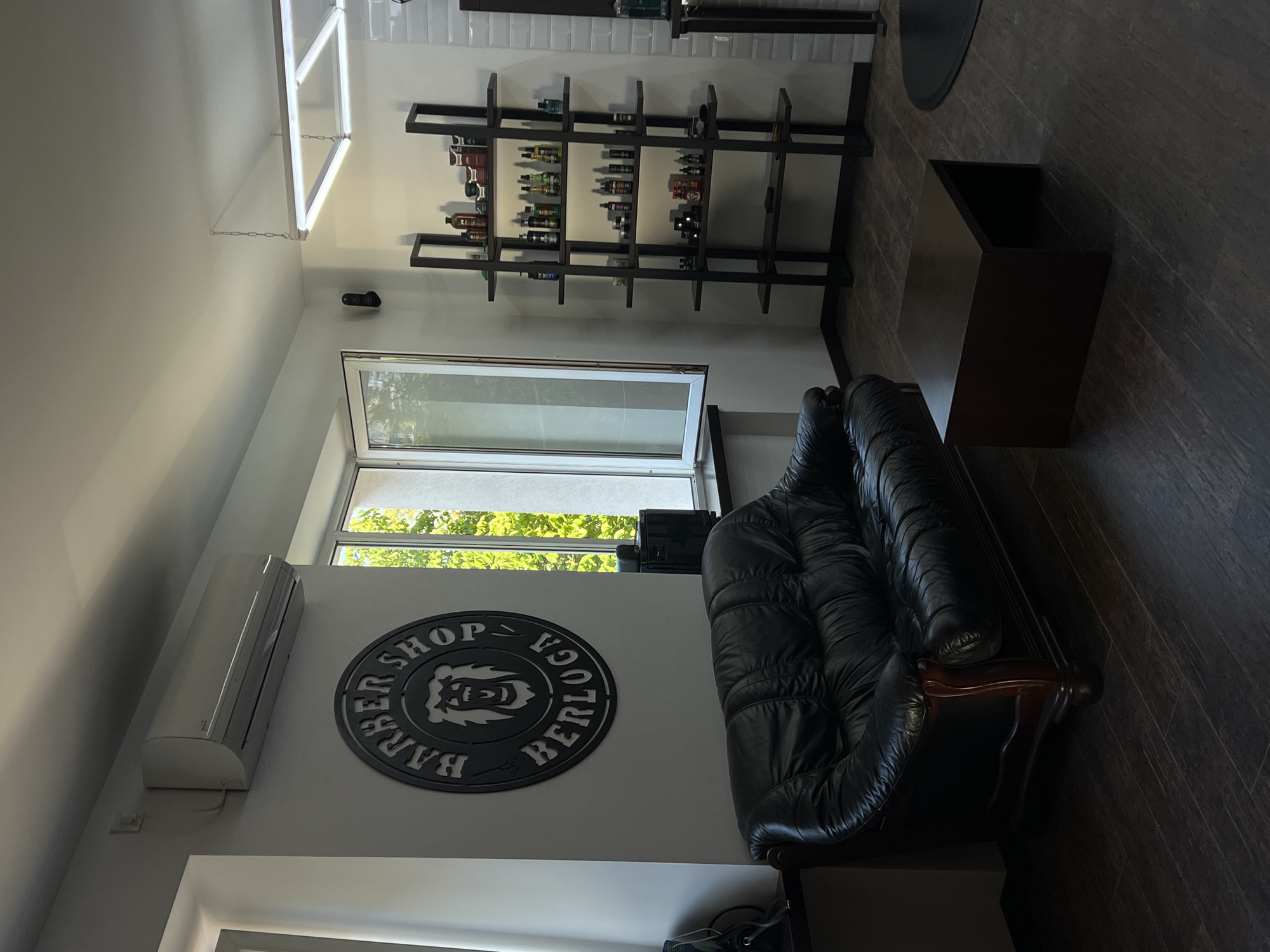 The image depicts the interior of a barber shop in Ternopil, Ukraine. The scene is captured with the camera angled downwards, giving a vertical perspective of the room. In the foreground, a black leather sofa with tufted detailing is prominent, positioned on a dark wood-grained floor. To the left of the sofa, an air conditioning unit is mounted high on a white wall. Below the air conditioner and to the right of it, a circular metal sign hangs on the wall, bearing the text "BARBER SHOP BERLOCA" in an arc around a stylized lion's head.
Further into the room, a large window allows natural light to illuminate the space, revealing lush green foliage outside, suggesting it is daytime. Above the window, a contemporary-style shelf unit is mounted on the wall. This unit displays numerous bottles, likely grooming products for hair and beards, arranged on several horizontal shelves. A dark rectangular object, possibly a coffee table or a storage unit, is placed on the floor to the right of the sofa. A suspended modern light fixture hangs from the ceiling, casting a bright light. The overall impression is a clean, modern, and well-lit barber shop.