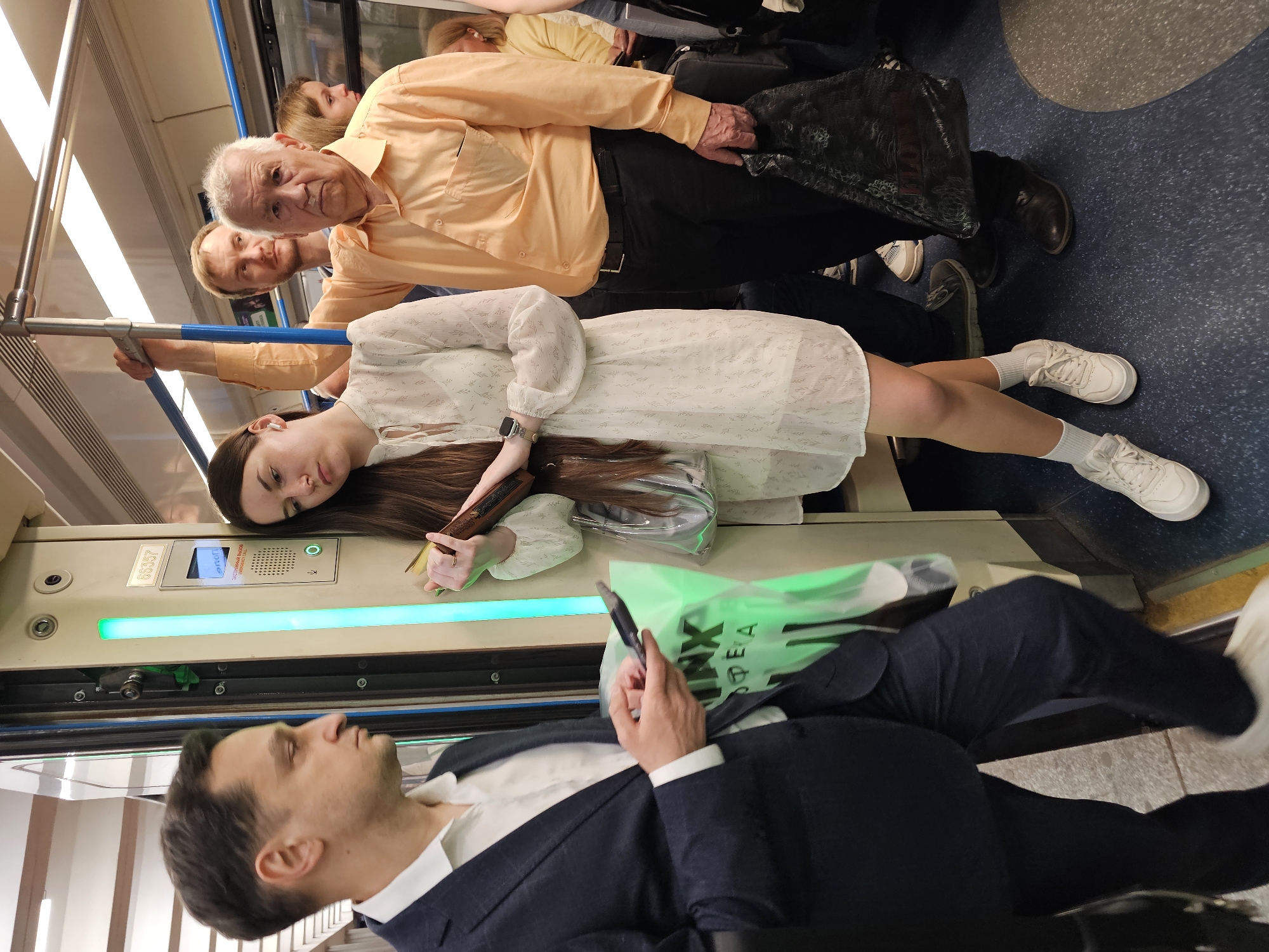 In a Moscow, Russia subway car, several people are present. A man in a dark suit is seated, looking down at a phone held in his hand. Next to him, a woman in a light-colored dress reclines, resting her head on the subway car wall. Behind her, an older man in a peach-colored shirt stands, holding onto a bar. Other people are visible in the background.