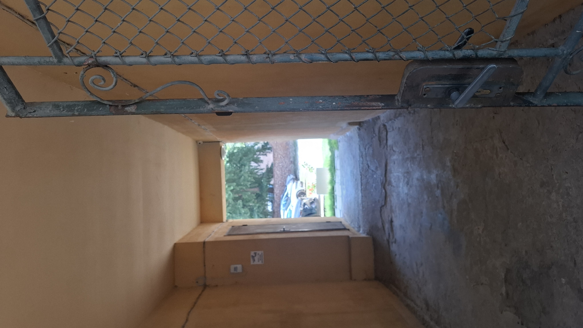 The image shows a downward view of a narrow outdoor passageway with two walls. The wall on the left is painted a light orange or tan color, while the wall on the right appears to be rough concrete or stone. At the top of the frame, a chain-link fence is visible, supported by a metal frame that appears to be rusted in places. A metal latch and lock mechanism is attached to this frame on the right side.
The passageway leads to an opening where sunlight is visible, suggesting daytime. Through the opening, trees and some greenery are visible, indicating an outdoor setting. In the distance, a white object, possibly a piece of outdoor furniture or equipment, can be seen. There is also a blue object, possibly a tarp or a bag, near the white object. The ground within the passageway is a rough, possibly dirt or gravel surface on the right, and a concrete or tiled surface on the left, leading to a set of stairs or a lower level.
On the left wall, there is a rectangular metal door or hatch set into a box-like structure, with what looks like a label or sign below it. A small white box, possibly an electrical outlet or intercom, is also visible on the left wall. The overall impression is one of a utilitarian, possibly neglected, outdoor space. The lighting suggests it is likely daytime, and the presence of greenery implies a temperate climate. The image does not depict any people or animals. There is no discernible text in the image.