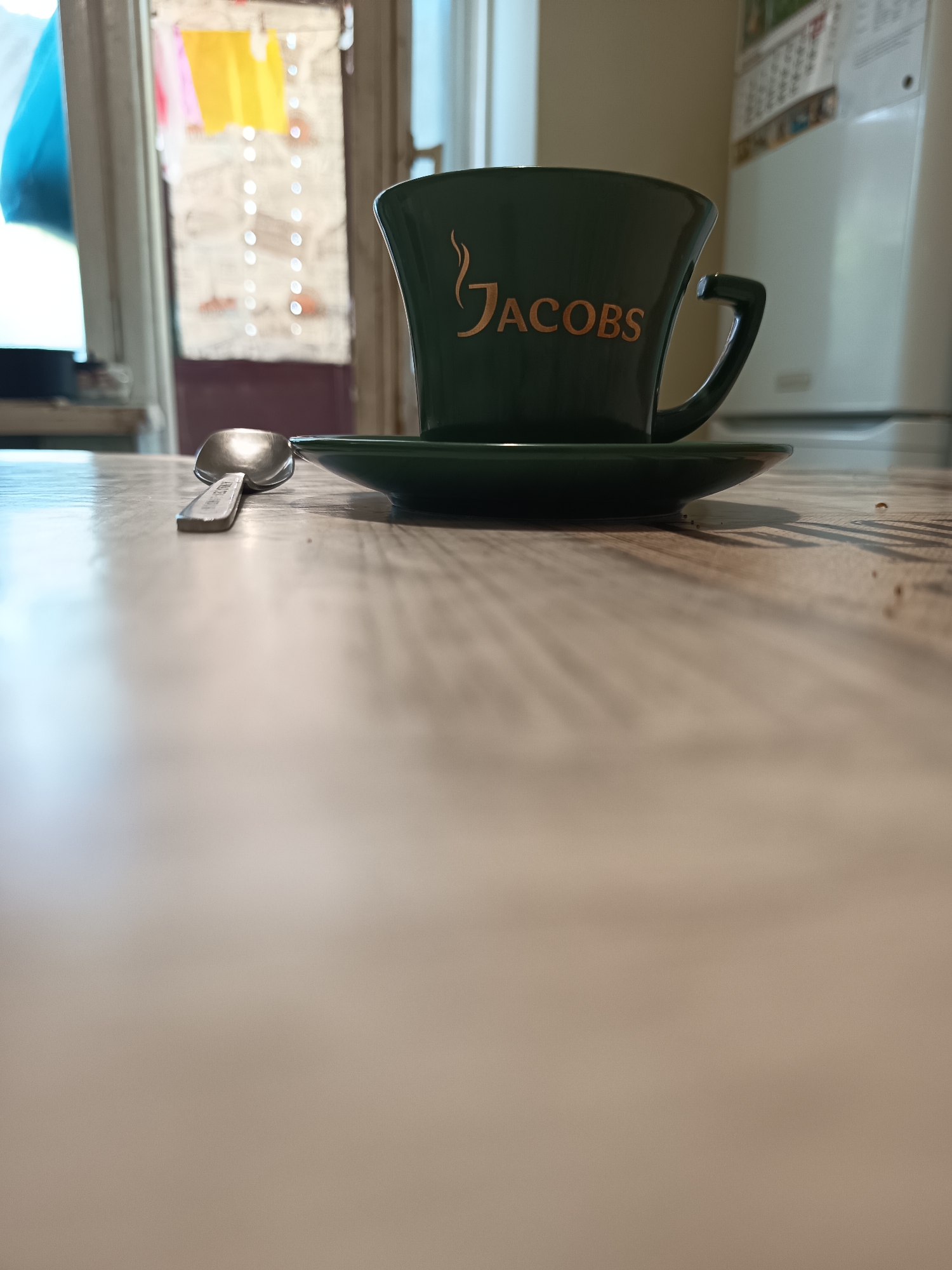 A close-up, low-angle shot shows a dark green Jacobs coffee cup and saucer on a light-colored wooden table. A silver spoon lies to the left of the cup. In the background, a doorway with laundry hanging is visible, along with part of a white appliance, possibly a refrigerator. The scene is indoors, and the lighting suggests it might be daytime. The overall setting appears to be a kitchen or dining area. There are no people visible in the image.