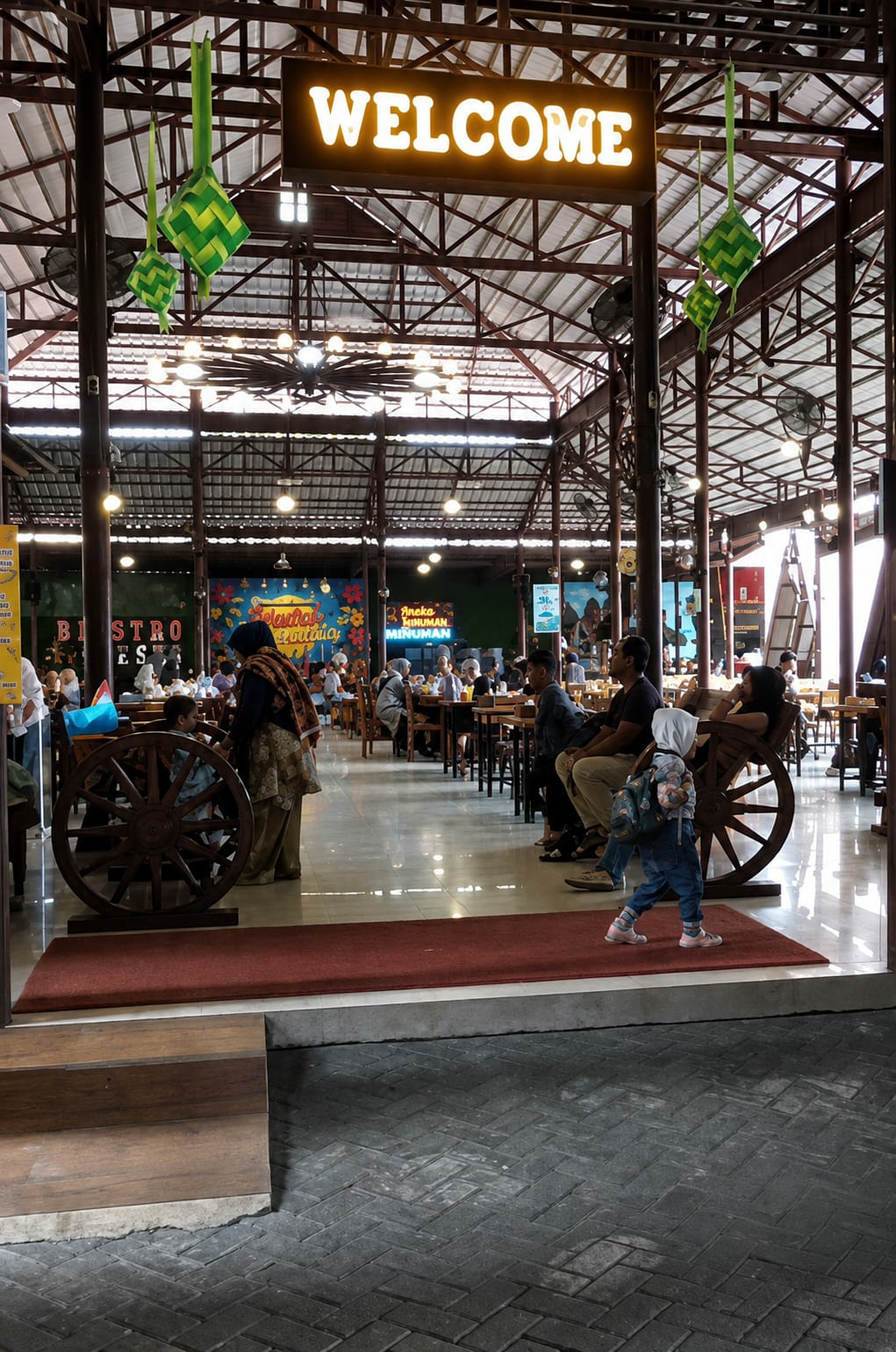 This image depicts the interior of a bustling restaurant or food court in the city of Binjai, Indonesia, with a prominent "WELCOME" sign illuminated above. The architecture features a high, exposed steel roof structure with numerous lights and fans. Ketupat decorations, traditional Southeast Asian rice cakes often associated with festive occasions, hang from the ceiling.
In the foreground, a young child wearing a hijab and carrying a backpack walks across a red carpeted area in front of a large wooden wagon wheel used as a decorative element. To the left of the wagon wheel, a woman in traditional attire is visible.
The middle ground shows a seating area with various tables and chairs occupied by diners. People are seen eating, conversing, and relaxing. In the background, more seating and serving areas are visible, along with decorative murals on the walls. One mural appears to have text in Indonesian, possibly indicating food or drink offerings. The floor is highly reflective, suggesting it is made of polished tile or stone. The overall atmosphere appears lively and social.