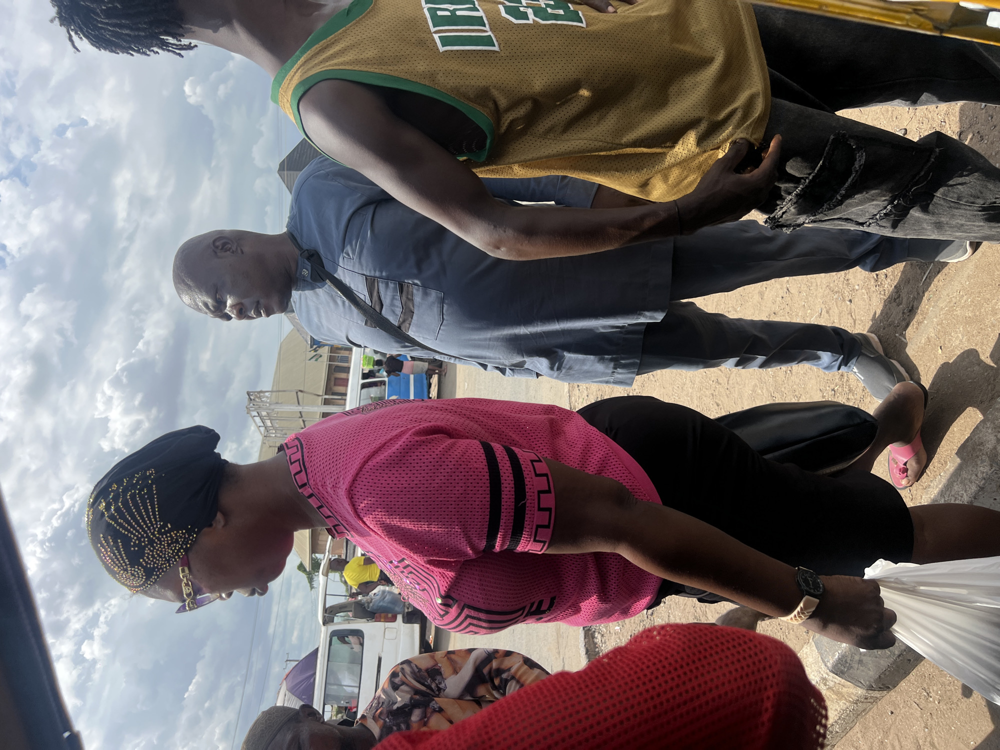 The image shows several people standing outdoors in Unknown, Unknown. A person with dreadlocks wears a yellow and green sports jersey. Another person in a blue button-down shirt and dark pants stands beside them. Further down, a woman in a pink mesh shirt, black pants, and a black head covering embellished with gold designs is holding a white plastic bag. Other individuals are partially visible in the background, and there are several white vans, indicating a possibly urban or commercial setting. The sky is overcast with scattered clouds.