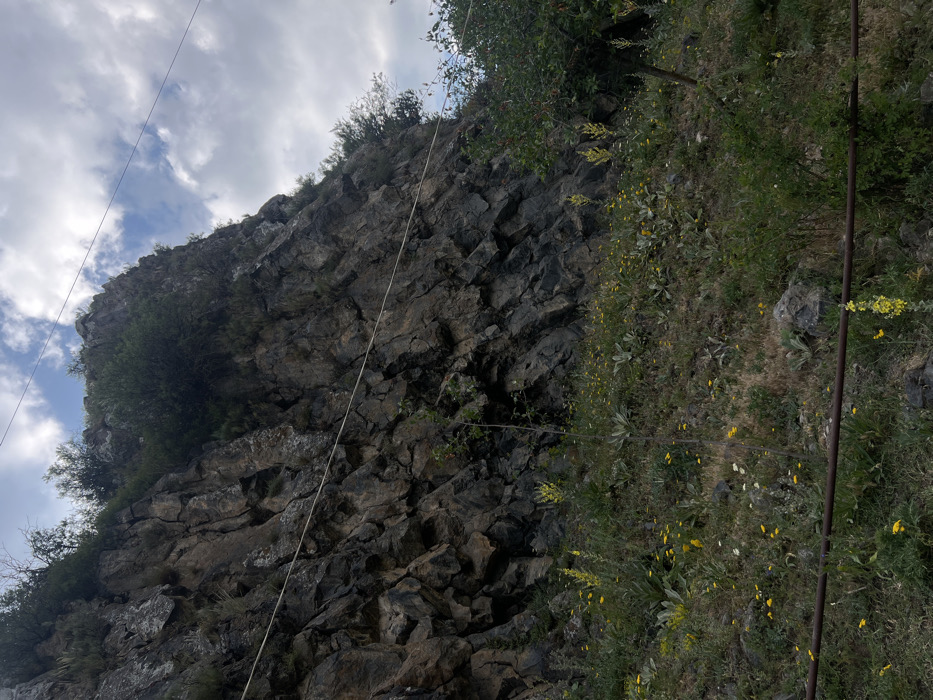 A steep rocky hillside dominates the image, with scattered green vegetation clinging to its surface. The rocks are dark and angular, forming a jagged texture. A thin white cable stretches across the scene, seemingly connecting the hillside to an unseen point. The sky is visible at the top of the frame, filled with wispy clouds. The vegetation appears more abundant on the right side of the image.