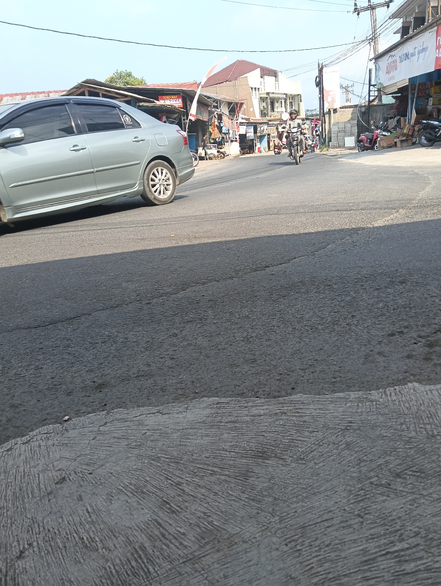 The image captures a street scene in Puspahiang, Indonesia, under a clear sky. In the foreground, a grey sedan is driving on an asphalt road. The car is positioned on the left side of the frame, with its rear tire visible. The road surface shows some wear and tear, with cracks and patches. To the right, a row of low-rise buildings lines the street, featuring a variety of signage, including what appears to be a "TOKO MAS" (gold shop) sign and another banner with text in Indonesian. Several motorcycles are parked in front of these establishments, and two individuals are riding a motorcycle on the road, heading away from the viewer. The buildings have tin roofs, and electrical wires are strung overhead. The overall impression is of a typical commercial street in a town.