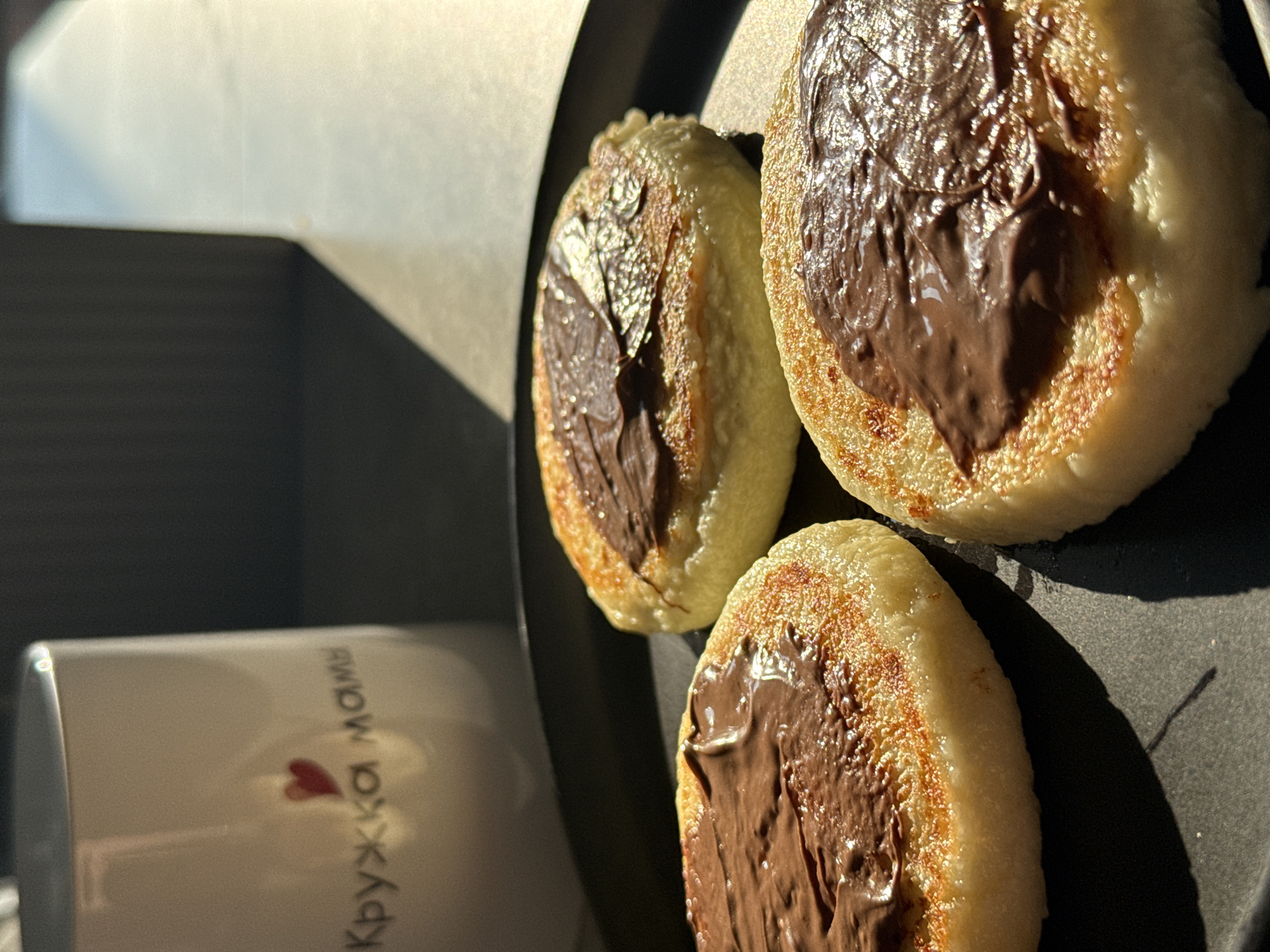 A close-up shot features three syrniki, a type of Russian cheese pancake, arranged on a black plate. Each syrnik is topped with a generous dollop of chocolate-hazelnut spread, appearing to be Nutella, which has been roughly applied, leaving visible texture. The syrniki themselves are golden brown on the edges, indicating they have been fried, and appear soft and fluffy in the center. In the background, out of focus, is a white ceramic mug with a red heart and Cyrillic text that reads "Кружка мамы" (Mom's Mug). The lighting suggests the scene is set indoors, possibly near a window, with sunlight casting shadows and highlighting the food. The overall impression is one of a simple, homemade breakfast or snack. This image was taken in Lyubertsy, Russia.