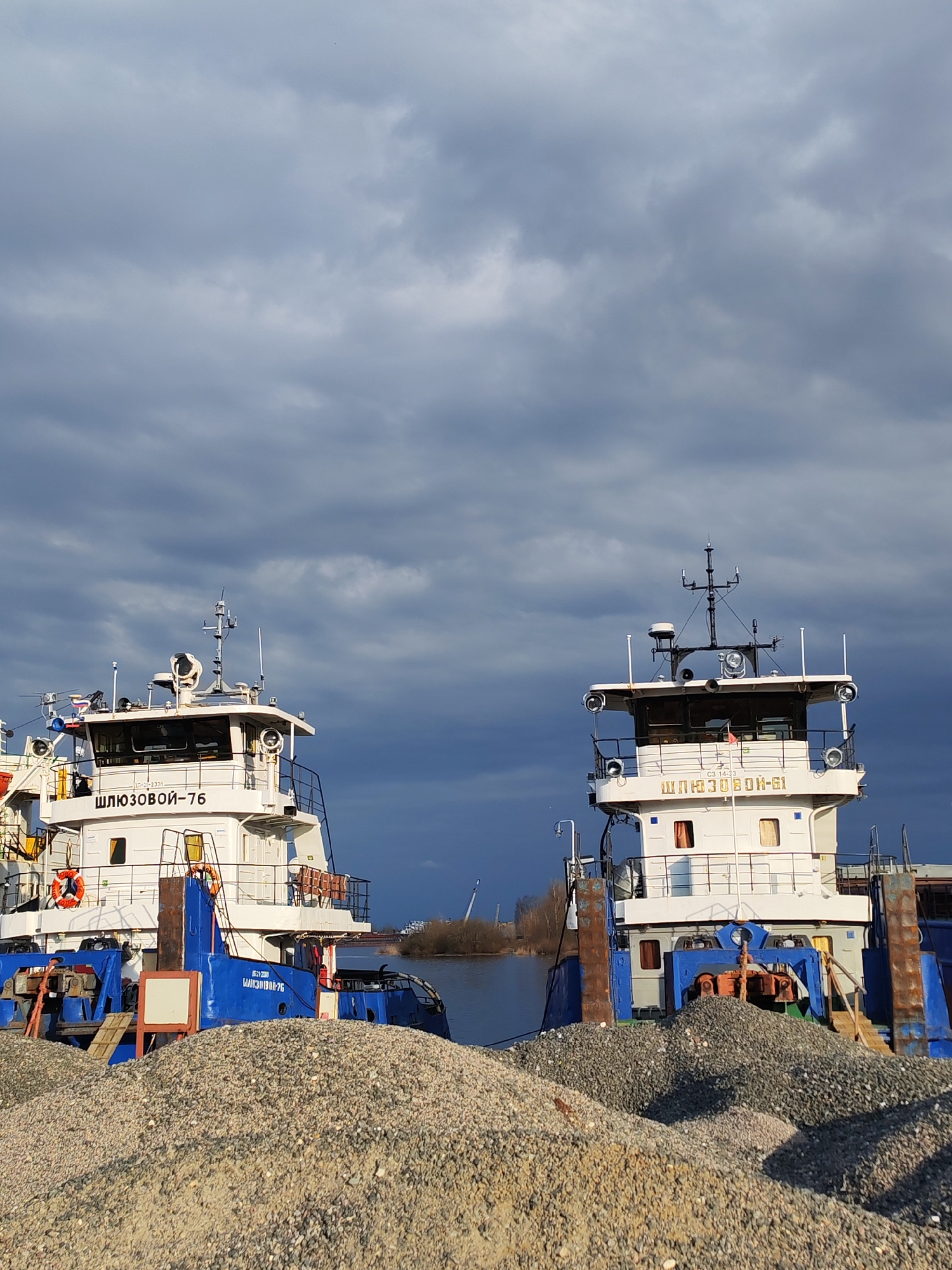 The image shows two ships docked next to each other in Rybinsk, Russia. The ship on the left is named "ШЛЮЗОВОЙ-76" (Shliuzovoy-76) and the one on the right is named "ШЛЮЗОВОЙ-61" (Shliuzovoy-61). Both ships are white with blue accents and have navigation equipment on their roofs. Piles of gravel are in front of the ships. The sky is filled with dark clouds. The river is visible in the background, with a bridge and some vegetation on the opposite shore.