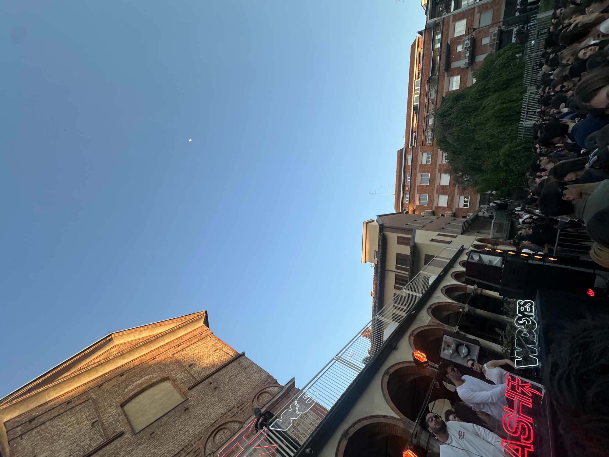 The image, captured in Milan, Italy, depicts an outdoor event taking place in an urban setting during the late afternoon or early evening. The perspective is tilted, showcasing a clear blue sky with a visible crescent moon and a small, distant flying object.
The scene is framed by two prominent buildings. On the left, an older, reddish-brown brick structure with a distinct roofline is bathed in warm, golden sunlight, highlighting its texture. On the right, a taller, multi-story residential building, also constructed of brick, features numerous windows and balconies.
In the mid-ground, beneath a series of architectural arches and a metal-railed walkway, a large crowd of people is gathered. They appear to be spectators, with many faces visible, predominantly looking towards the center of the frame. In the foreground, below the arches, a stage or DJ booth is set up with large speakers and illuminated elements. Several individuals, likely performers or DJs, are positioned behind the equipment. One person wears a white shirt with the letters "GD" visible.
Notable details include the clear weather and the low sun angle, which casts long shadows and provides a golden hue to the brickwork, suggesting the time is either late afternoon or early evening. Two distinct neon signs are prominently displayed on the stage structure: "WASHIF" in bright red lettering and "WDSES" in white. The blend of historic and modern urban architecture provides a rich backdrop for the gathering.