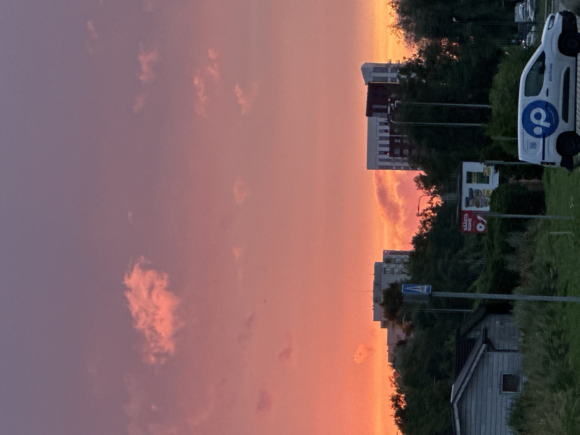 This is an outdoor, vertical, medium-angle, eye-level photograph of a suburban area at sunset, likely in Tallinn, Estonia, given the presence of a van with the logo "Parkir" which is a service operating in Estonia. The sky is filled with vibrant hues of pink, orange, and purple, with a few wispy clouds scattered throughout. In the foreground, on the left, there is a small, gray, wooden house with a dark roof and a single window. Adjacent to it is a patch of overgrown grass and vegetation. A gray metal pole with a blue and white pedestrian crossing sign stands in the middle of the frame. Behind the house and pole, there is a dense area of green trees and shrubs. Further back, a modern white and dark-paneled apartment building with many windows is visible, towering above the trees. To the right of this building, another, older, gray apartment building is partially visible. In the lower right corner, a white panel van with a blue logo featuring a stylized "p" and the word "Parkir" written in blue lettering is parked on a road. The overall scene depicts a quiet evening in a residential area with an impressive sunset.