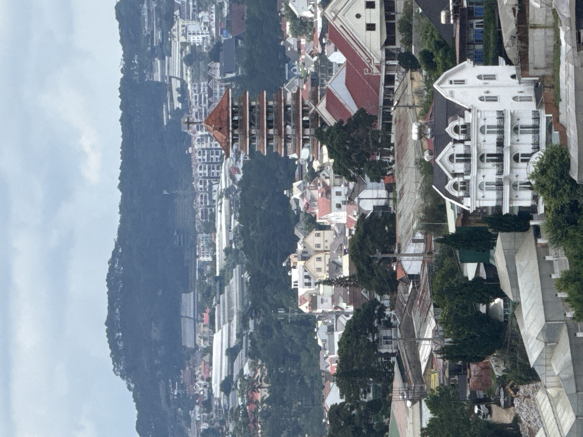 The image captures a panoramic view of Đà Lạt, a city in the Lâm Viên highlands of Vietnam. In the foreground, a cluster of buildings with red tile roofs are visible, interspersed with lush green trees. A distinctive white building with arched windows and balconies stands out among them. Further back, a multi-tiered pagoda-like structure with a red roof dominates the mid-ground. Beyond this, the city sprawls across rolling hills, characterized by a mix of residential buildings, some with white facades and red roofs, and numerous greenhouses with translucent coverings, indicating agricultural activity. The backdrop is a dark, forested mountain range under a cloudy sky. The scene depicts a densely populated urban area integrated with natural landscapes and agricultural infrastructure.