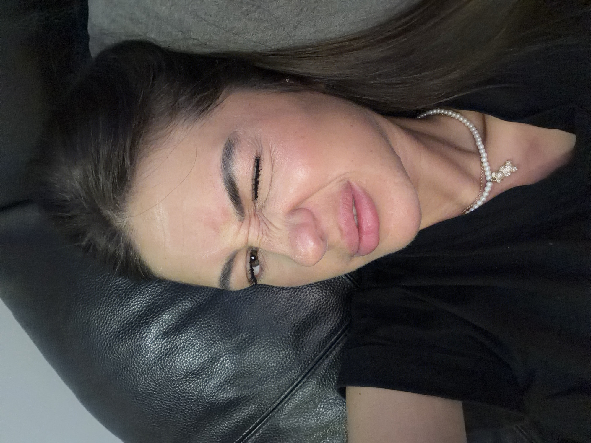 The image shows a close-up, waist-up, selfie of a young woman. She has long, dark brown hair and is wearing a black t-shirt and a pearl necklace with a teddy bear pendant. The woman is winking and has her nose scrunched up, suggesting she is making a funny face. She is lying down, with her head resting on a dark, textured surface, possibly a couch or a pillow. A grey surface is visible behind her head. The lighting is warm and slightly diffused. The image was taken indoors. The woman is of European descent.