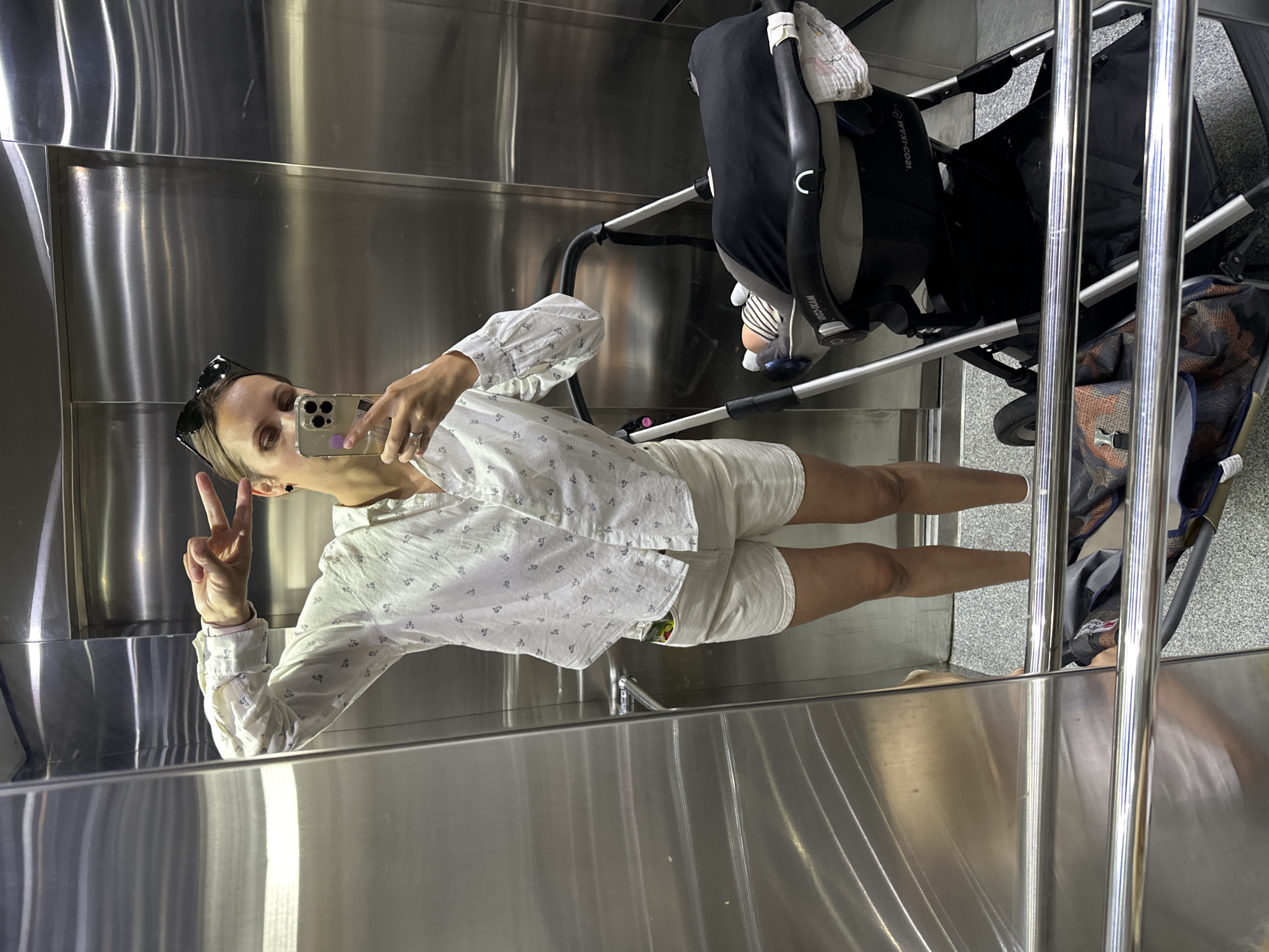 The image shows a woman taking a selfie in an elevator. She is wearing a white button-down shirt with a small pattern and white shorts. She is holding up a peace sign with one hand while holding a smartphone in the other. She has sunglasses resting on her head. Next to her is a stroller containing a baby. The elevator interior is metallic, possibly stainless steel. The location is not specified in the prompt, so I cannot provide a location in Fethiye, Turkey.