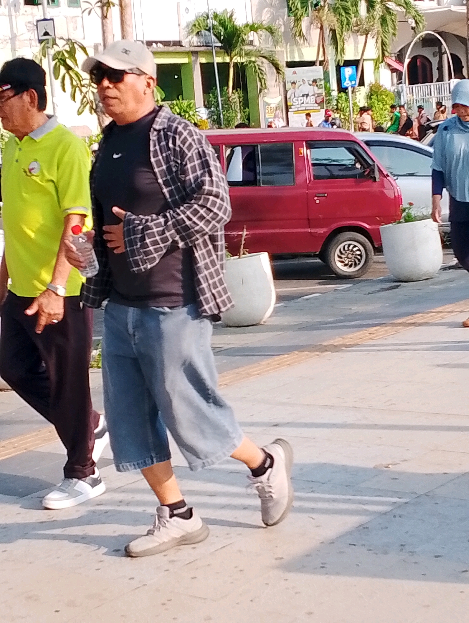 The image depicts a street scene in Probolinggo, Indonesia. In the foreground, two men are walking on a sidewalk. The man on the left wears a bright green polo shirt and dark trousers. He is holding a bottle of water. The man on the right wears a black t-shirt, a plaid open shirt, and light blue denim shorts. He is also wearing a light-colored baseball cap and sunglasses. Behind them, a red van is parked on the street, and two large, light-colored planters are visible on the sidewalk. In the background, buildings and palm trees can be seen. The overall impression is of a casual daytime scene in an urban environment.