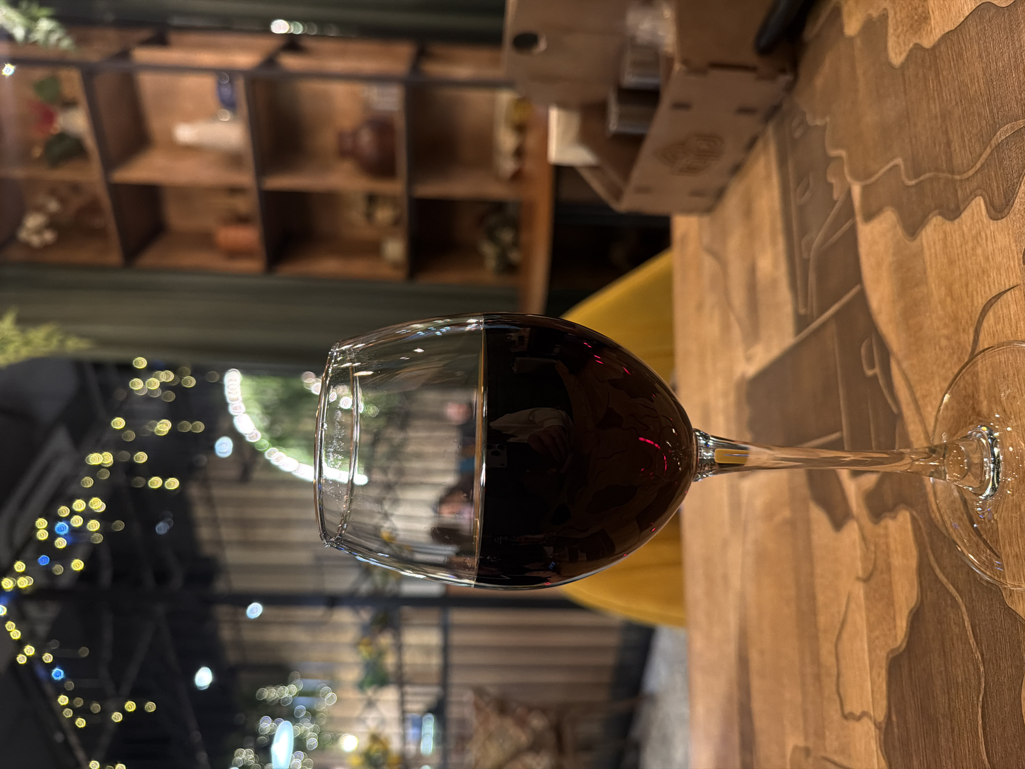 A close-up, slightly blurry, high-angle shot depicts a partially filled wine glass resting on a light brown wooden table. The liquid inside the glass appears to be red wine. The table has a patterned surface with darker brown carvings or engravings. Behind the glass, there is a blurred background featuring string lights and greenery. In the upper left of the frame, blurry wooden shelves with assorted objects are visible. A yellow chair is behind the wine glass. There is a box of wooden decor in the background. The location is Izhevsk, Russia. The focus is on the wine glass.