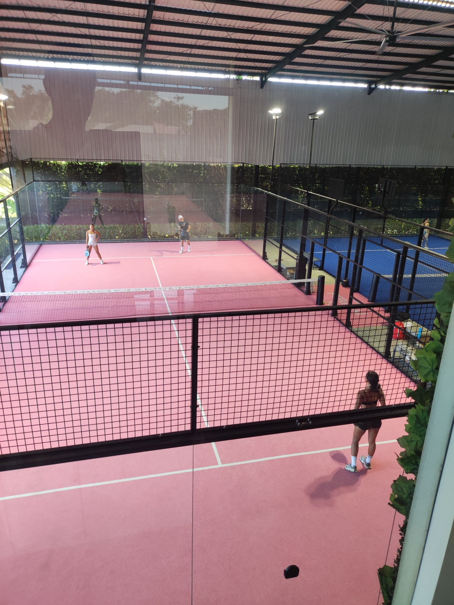 The image captures a high-angle view of a padel tennis facility in North Kuta, Indonesia. The indoor facility is equipped with at least two padel courts, one pink and one blue, each enclosed by black metal mesh fencing.
On the pink court further from the camera, two individuals are engaged in a game of padel tennis. One person wears a white outfit, and the other wears a grey or black top with shorts.
On the pink court in the foreground, a woman wearing a dark sports bra and shorts stands on the court, facing away from the camera, towards the net. A small, dark object rests on the court near her.
The facility has a high roof with metal beams and artificial lighting. Greenery is visible outside the court enclosures, suggesting an open-air or partially open-air design.