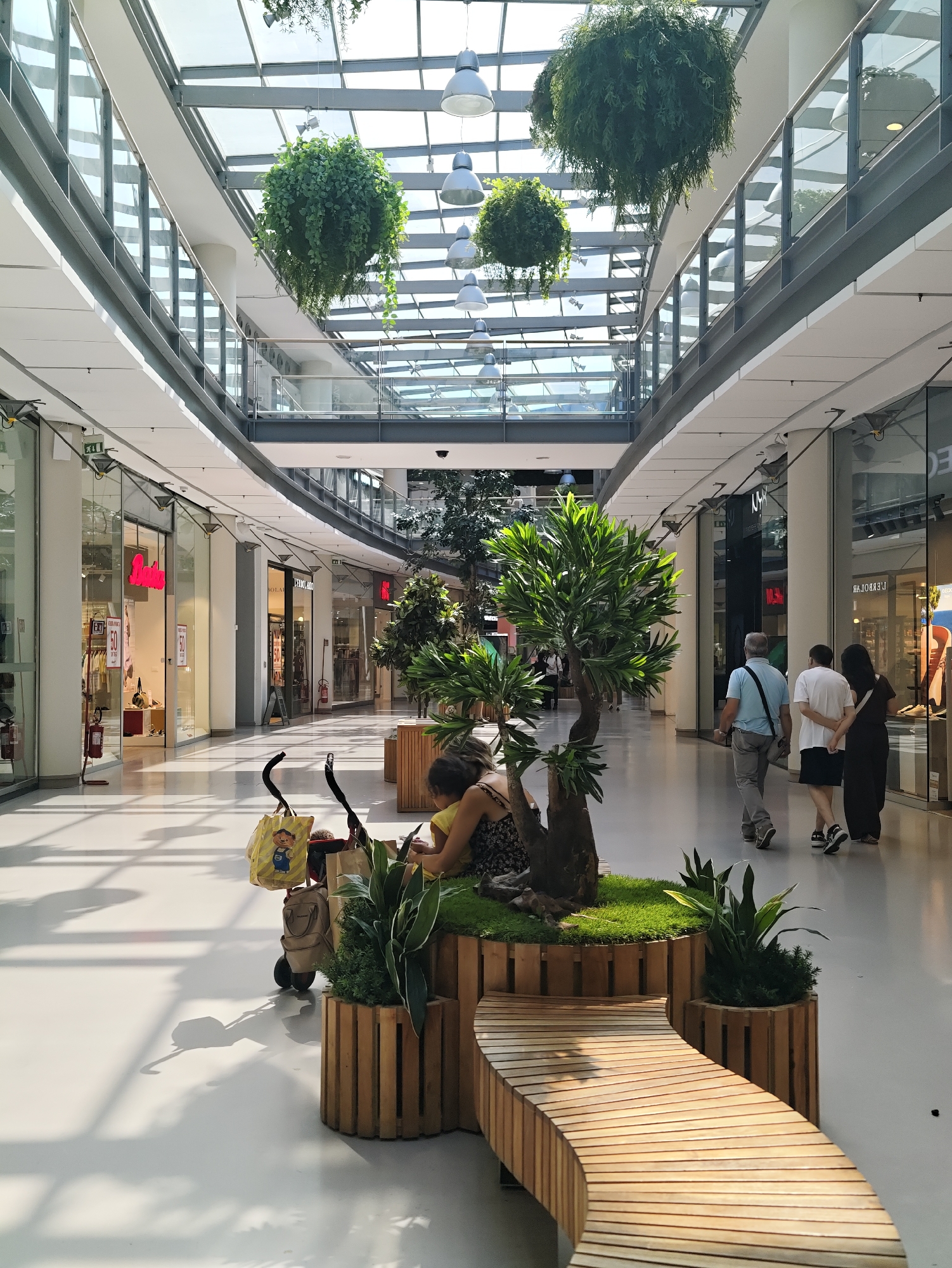 The image shows the interior of a modern shopping mall in Nola, Italy, with a glass ceiling that allows natural light to enter. Hanging plants are suspended from the ceiling, adding a touch of greenery to the environment. In the foreground, a woman with dark hair is sitting on a wooden bench, facing away from the camera, with her back to a stroller. The stroller contains several shopping bags. Further down the mall, a man in a light blue shirt and grey pants, a young person in a white t-shirt and black shorts, and a woman in a dark dress are walking away from the camera. Shops with glass storefronts line both sides of the mall, and display items and signage can be seen. The flooring is a light-colored, smooth surface, reflecting the light. The overall atmosphere is clean, bright, and contemporary.