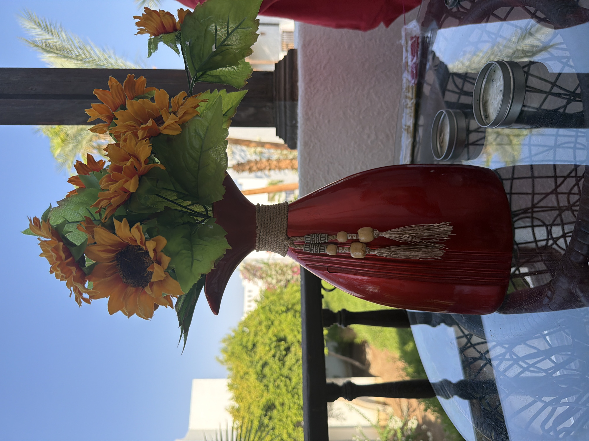 The image depicts a still life scene on a glass-top table, likely outdoors given the background. In the foreground, a glossy red vase filled with artificial sunflowers and green leaves stands prominently. The vase is adorned with a rope detail around its neck, from which hang two beaded tassels. To the right of the vase, two small, round metal tins are visible on the table. The background reveals a railing with vertical posts, beyond which is lush green foliage and a building. In the far background, a palm tree and a clear blue sky are visible, suggesting a warm, possibly tropical or subtropical, climate. The scene is set in Sharm El Sheikh, Egypt, a location known for its resorts and clear skies. There are no people or animals present in the image.