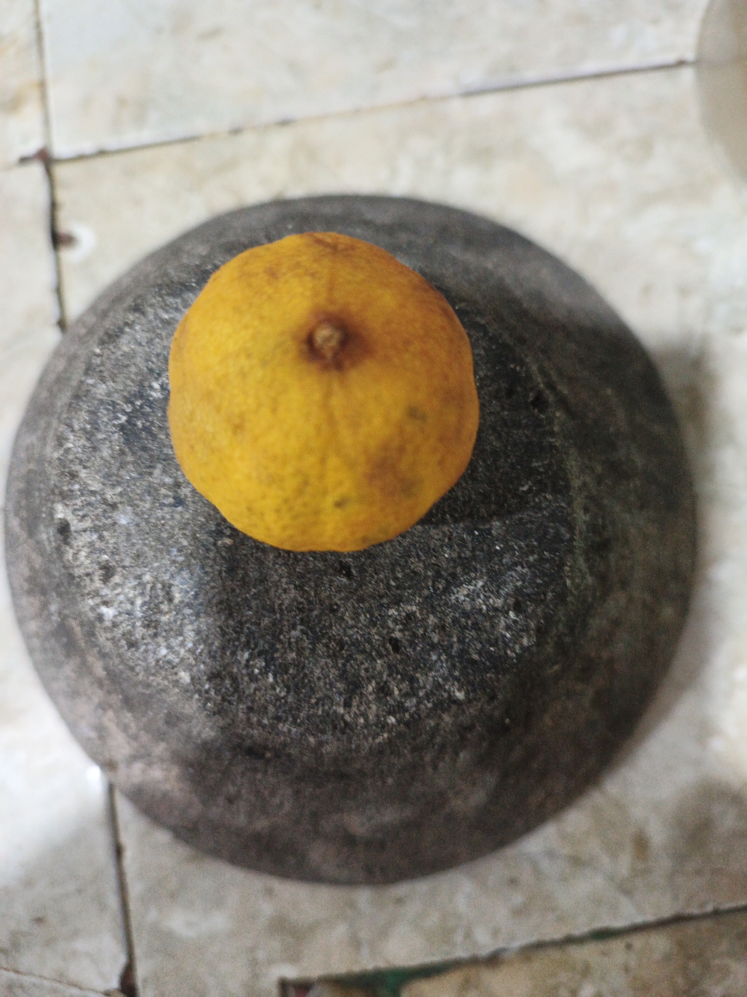 An overhead shot shows a single orange resting on top of a mortar. The mortar is made of dark, rough stone and is placed on a beige tiled surface. The orange is roughly spherical and vibrantly colored. The scene appears to be indoors, given the tiled floor and artificial lighting. There are no people or animals present.