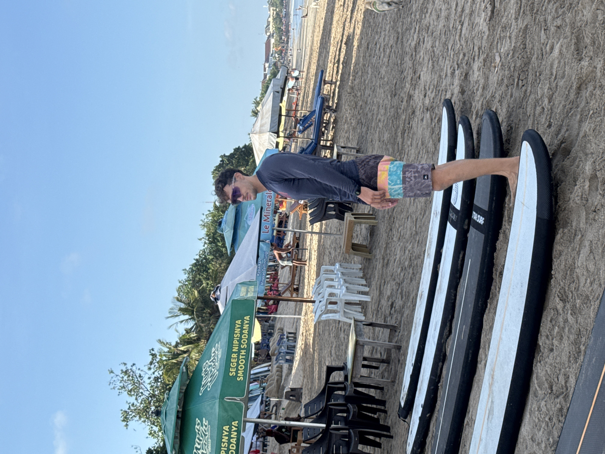 The image shows a man standing next to a row of surfboards on a beach in Kuta, Indonesia. The man is wearing sunglasses, a long-sleeved shirt, and patterned shorts. He is standing facing the ocean. To the left of the man is an outdoor dining area with umbrellas bearing the logos of "Le Mineral" and "Nipis." In the background, the ocean and sky are visible. The beach appears to be sandy.